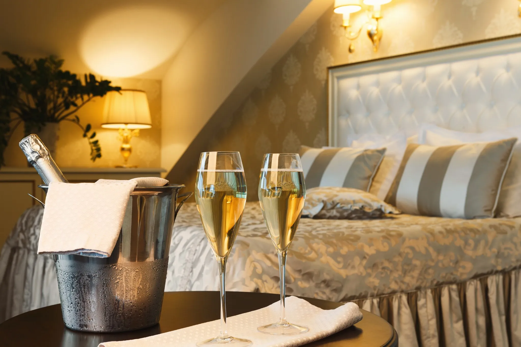 Sektkühler und zwei gefüllte Gläser auf einem Tisch vor einem Hotelbett mit gepolstertem Kopfteil