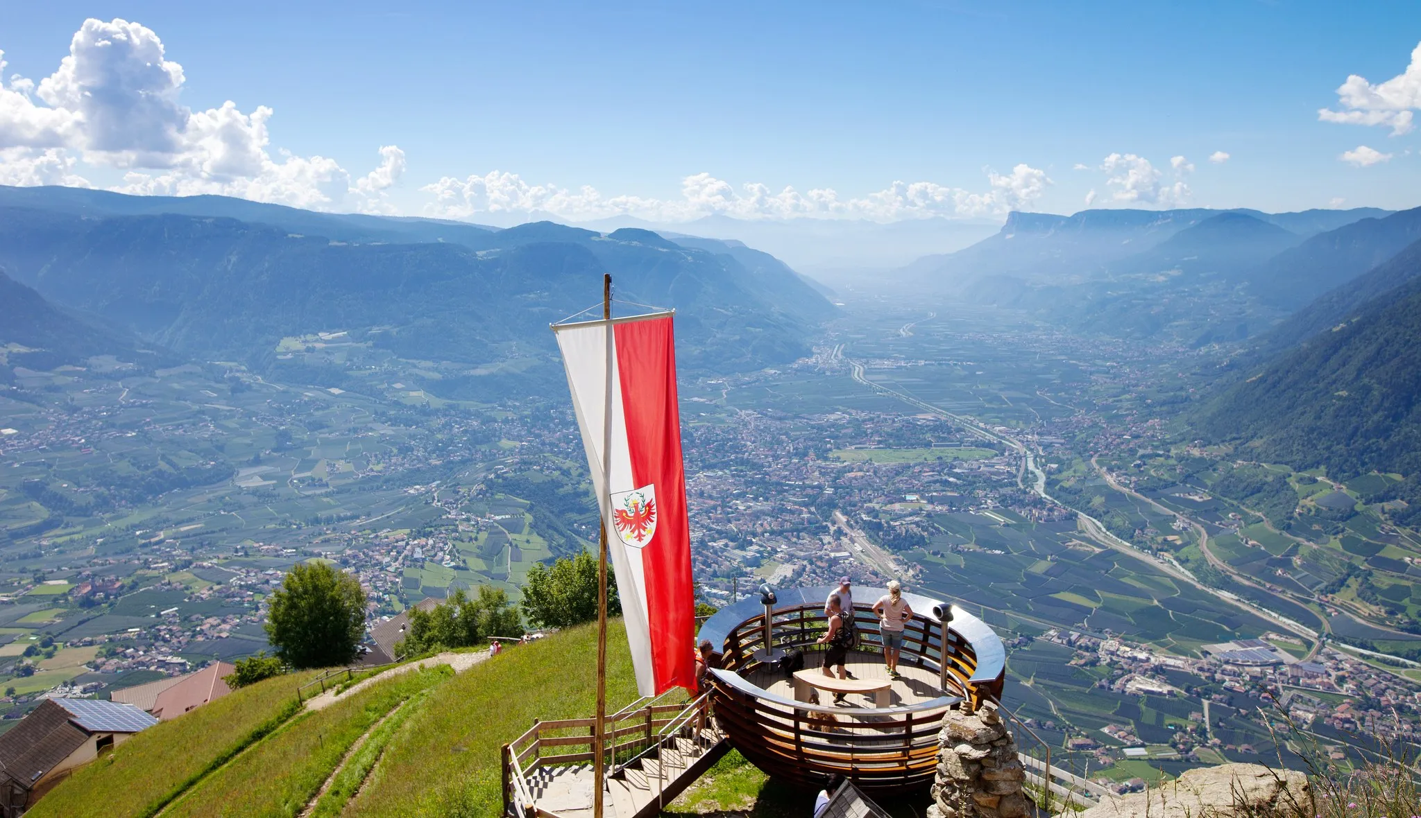 Aussichtsplattform mit Südtiroler Fahne hoch über dem Etschtal, im Talgrund die Stadt Meran