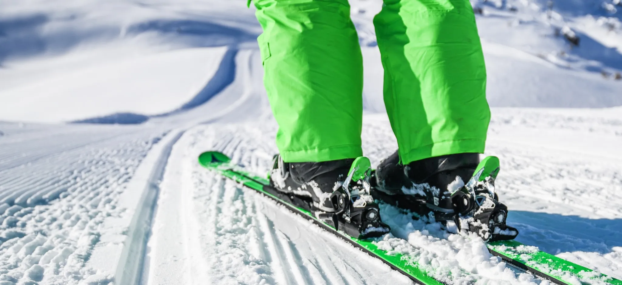 Beine in grüner Skihose auf Skiern am Rand einer frisch präparierten Piste mit sichtbaren Pistenraupenspuren
