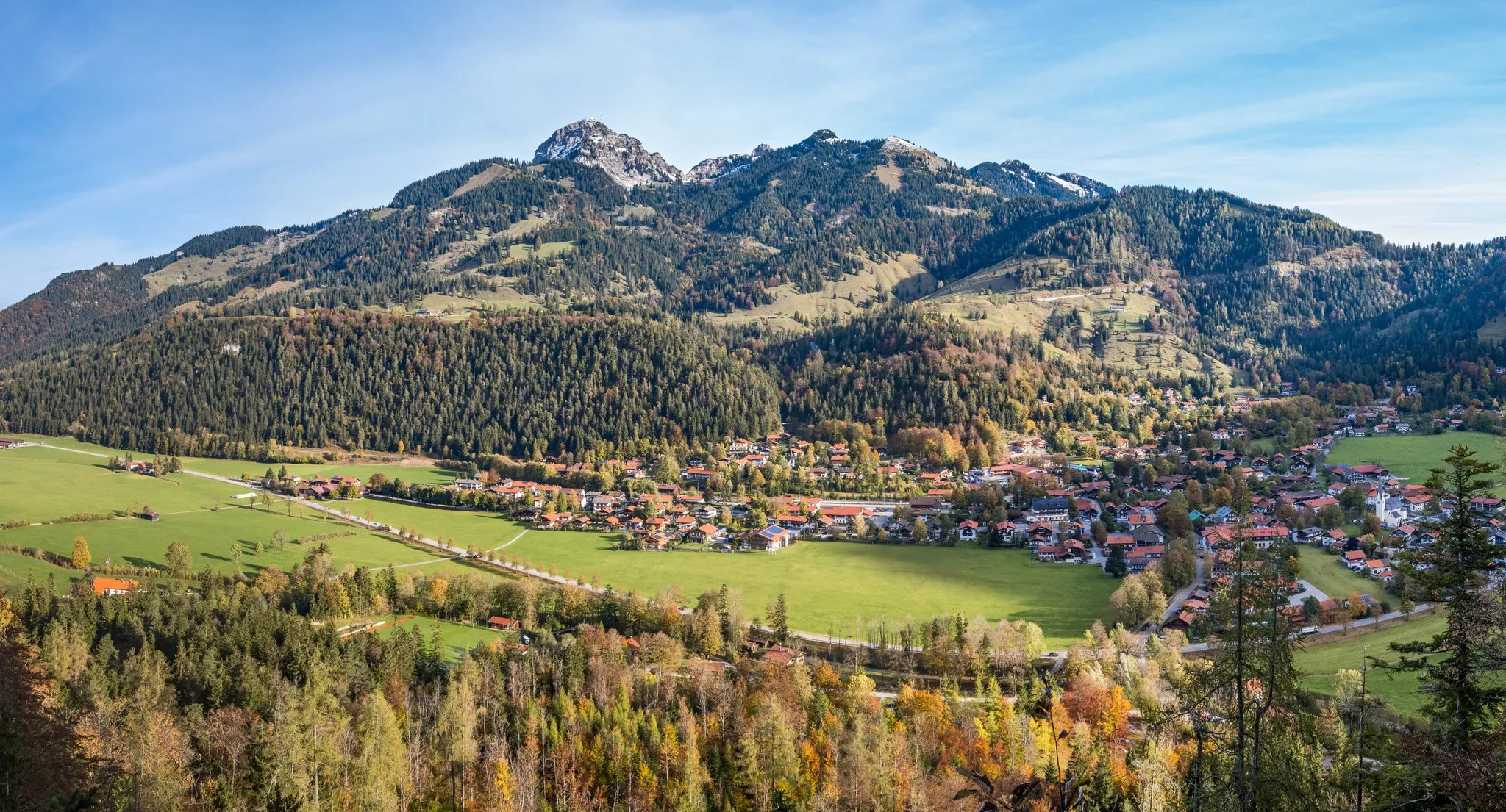 Blick über ein Alpendorf im Talgrund auf einen bewaldeten Bergrücken mit felsigem Gipfel im Herbst