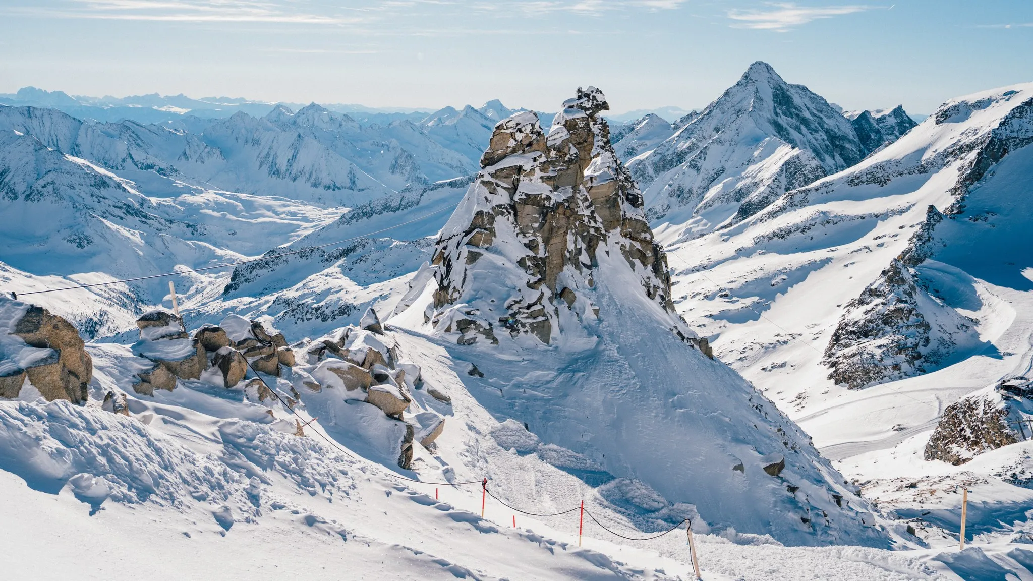 Verschneite Gletscherlandschaft mit freistehender Felsnadel und markierter Piste vor gestaffelten Alpengipfeln