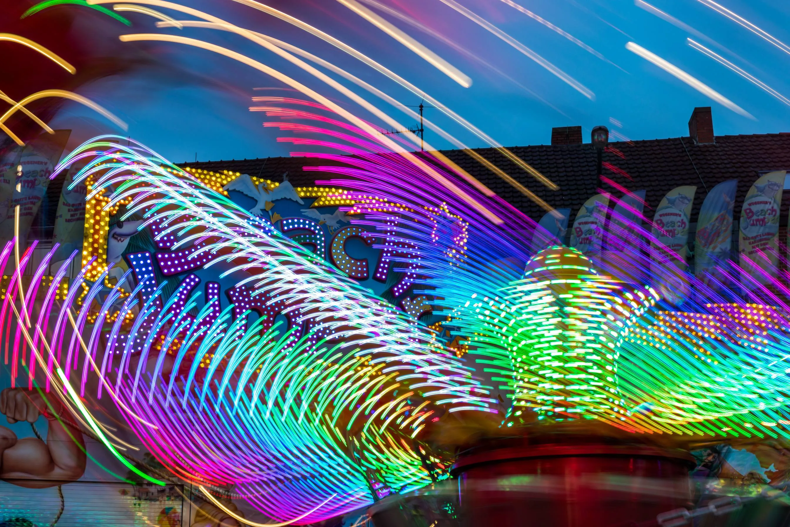 Lichtspuren eines Fahrgeschäfts in der Abenddämmerung, bunte Leuchtbögen über einem Kirmesplatz