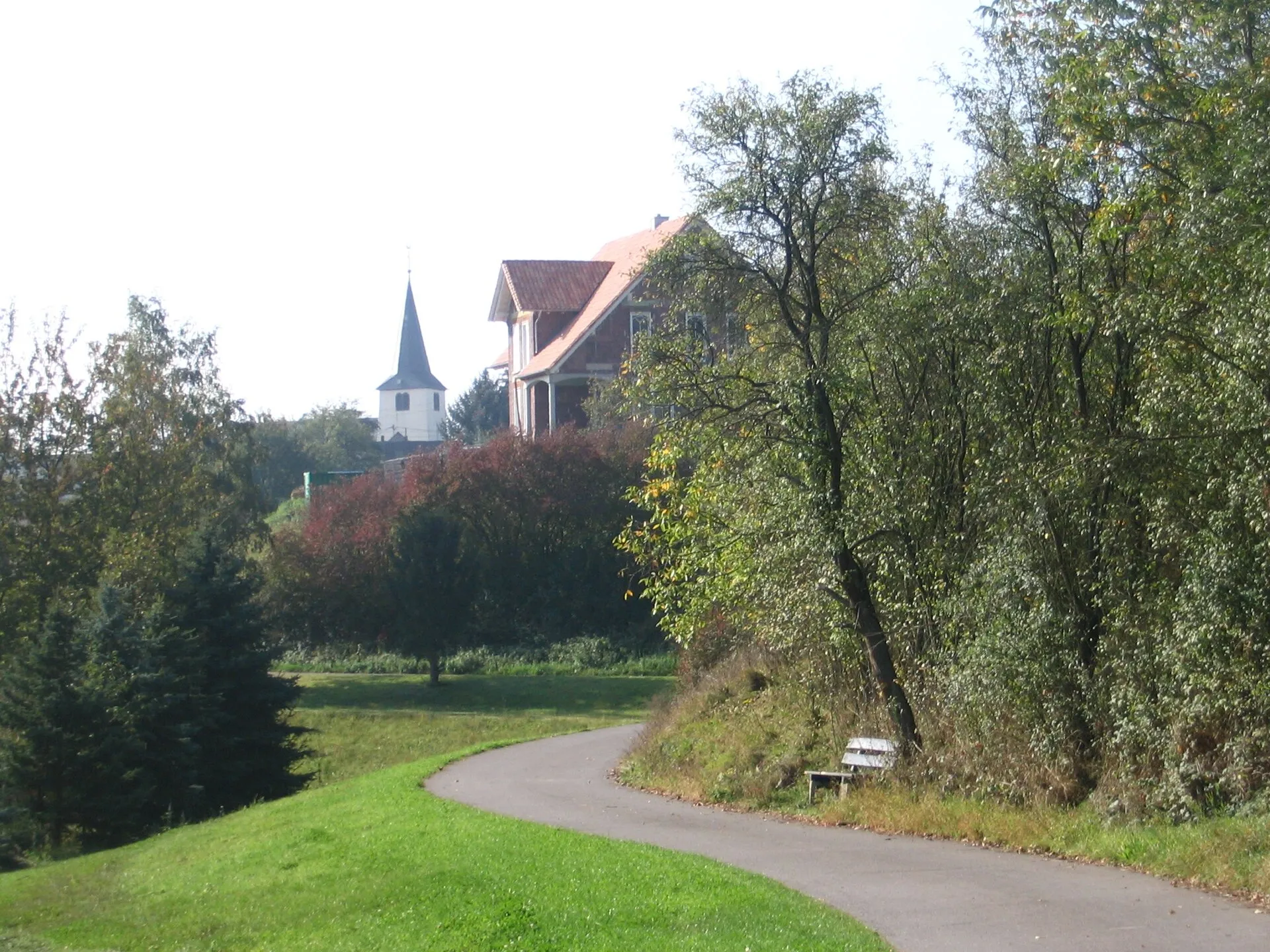 Asphaltierter Radweg, der an einer Wiese entlang zu einem Dorf mit spitzem Kirchturm ansteigt