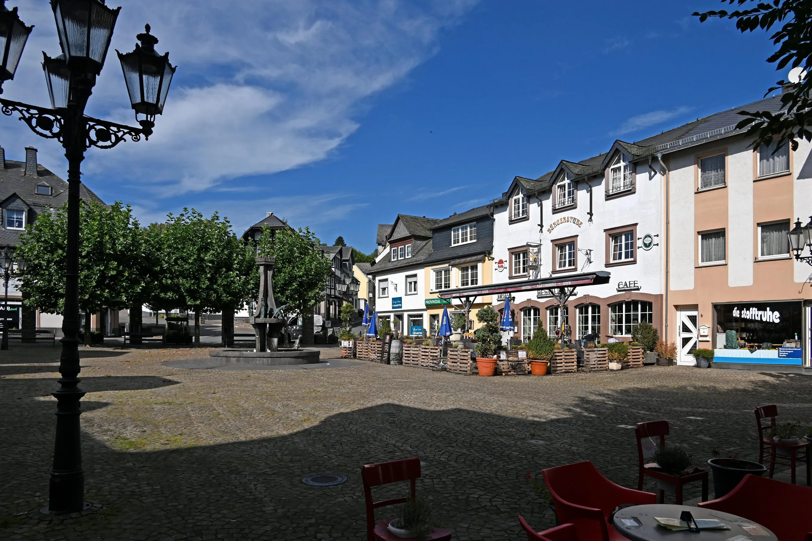 Alter Postplatz in Ulmen mit Brunnen, Lindenreihe und Fachwerkhäusern, davor leere Caféstühle