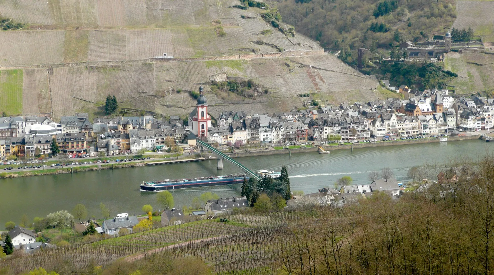 Blick über die Mosel auf Zell mit Kirchturm, Uferbebauung, Brücke und einem Frachtschiff, ringsum Weinberge