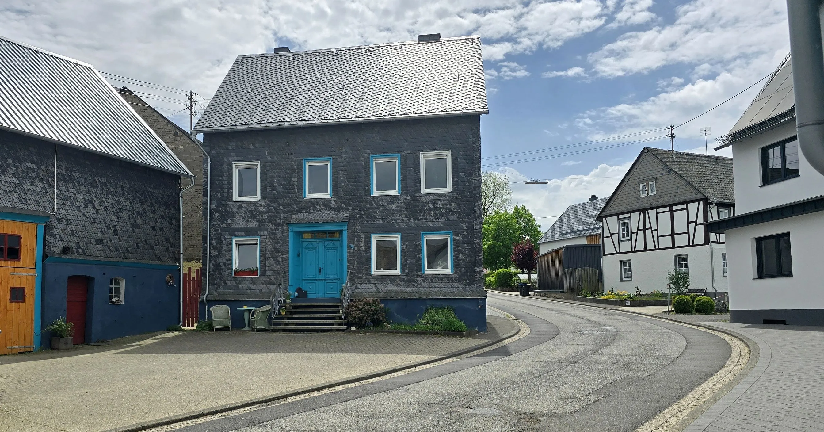 Schiefergedecktes Wohnhaus mit blauer Haustür an einer Straßengabelung in Blankenrath, rechts ein Fachwerkhaus