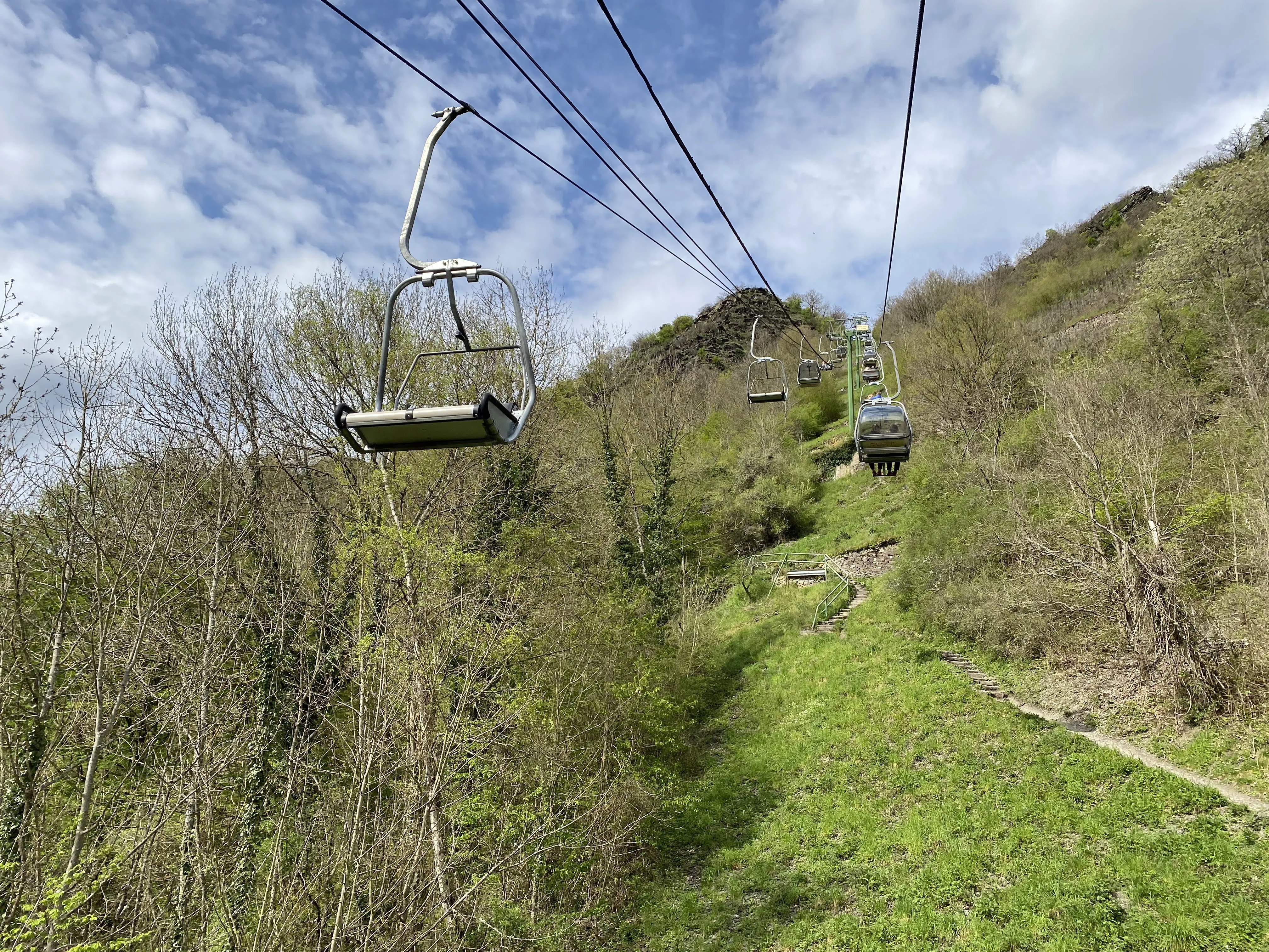 Sesselbahn über einem grünen Hang, daneben ein steiler Treppenweg bergauf, im Hintergrund bewaldete Höhen