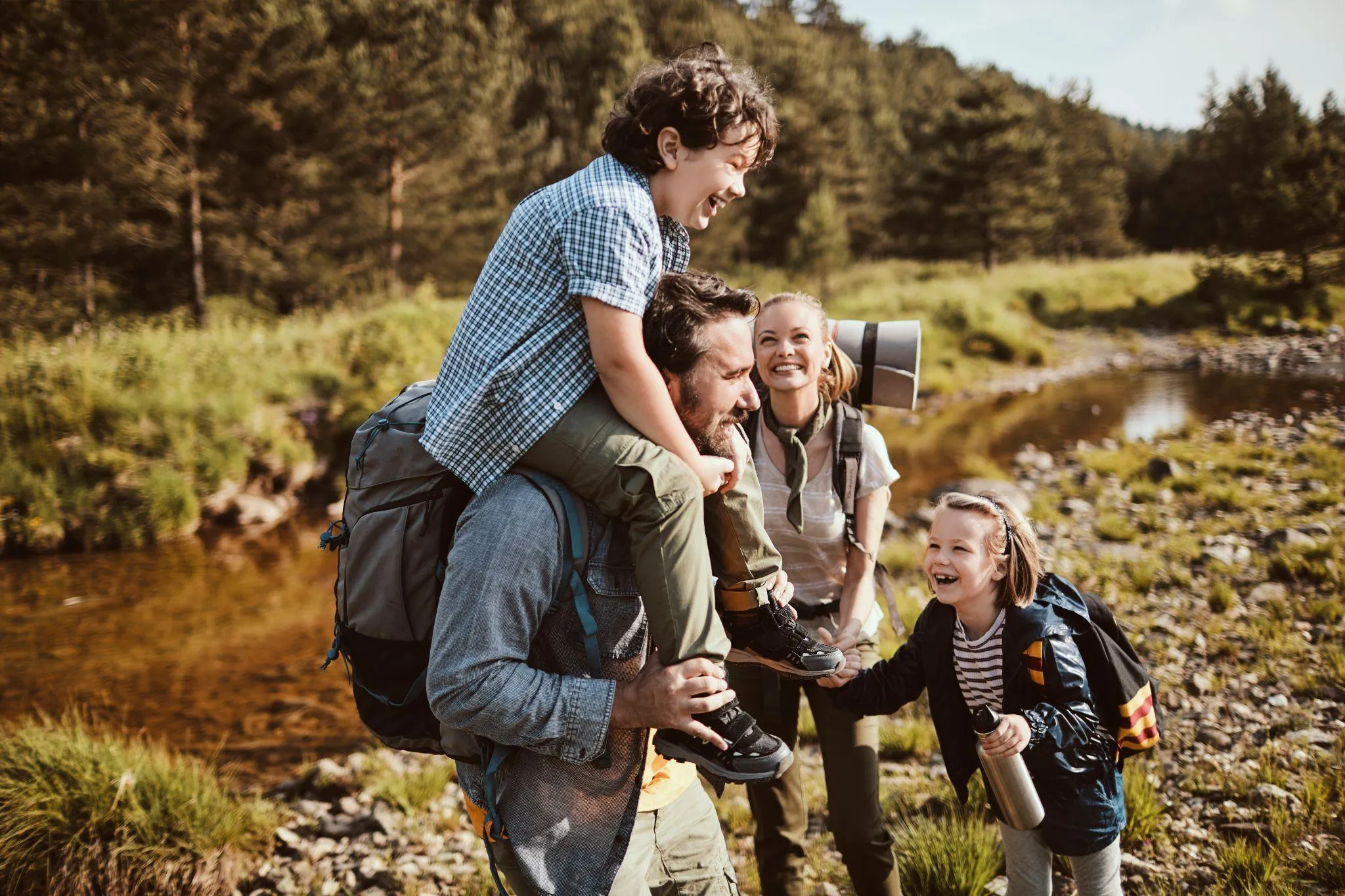 Familie auf Wanderung an einem Bach, der Vater trägt den Sohn auf den Schultern, die Tochter hält eine Trinkflasche