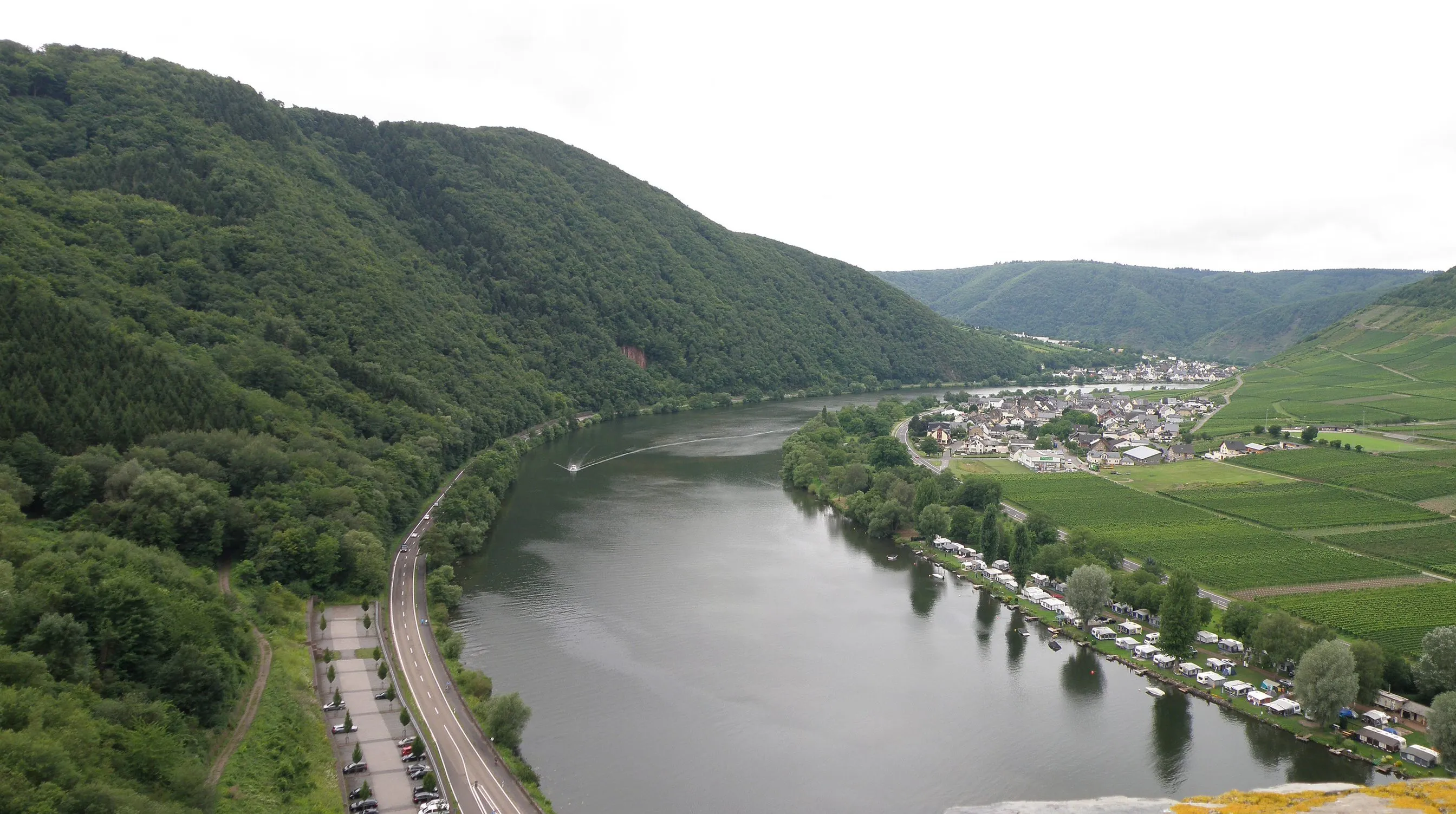 Blick über die Mosel: links eine Uferstraße am bewaldeten Hang, rechts Poltersdorf mit Weinbergen und Campingplatz
