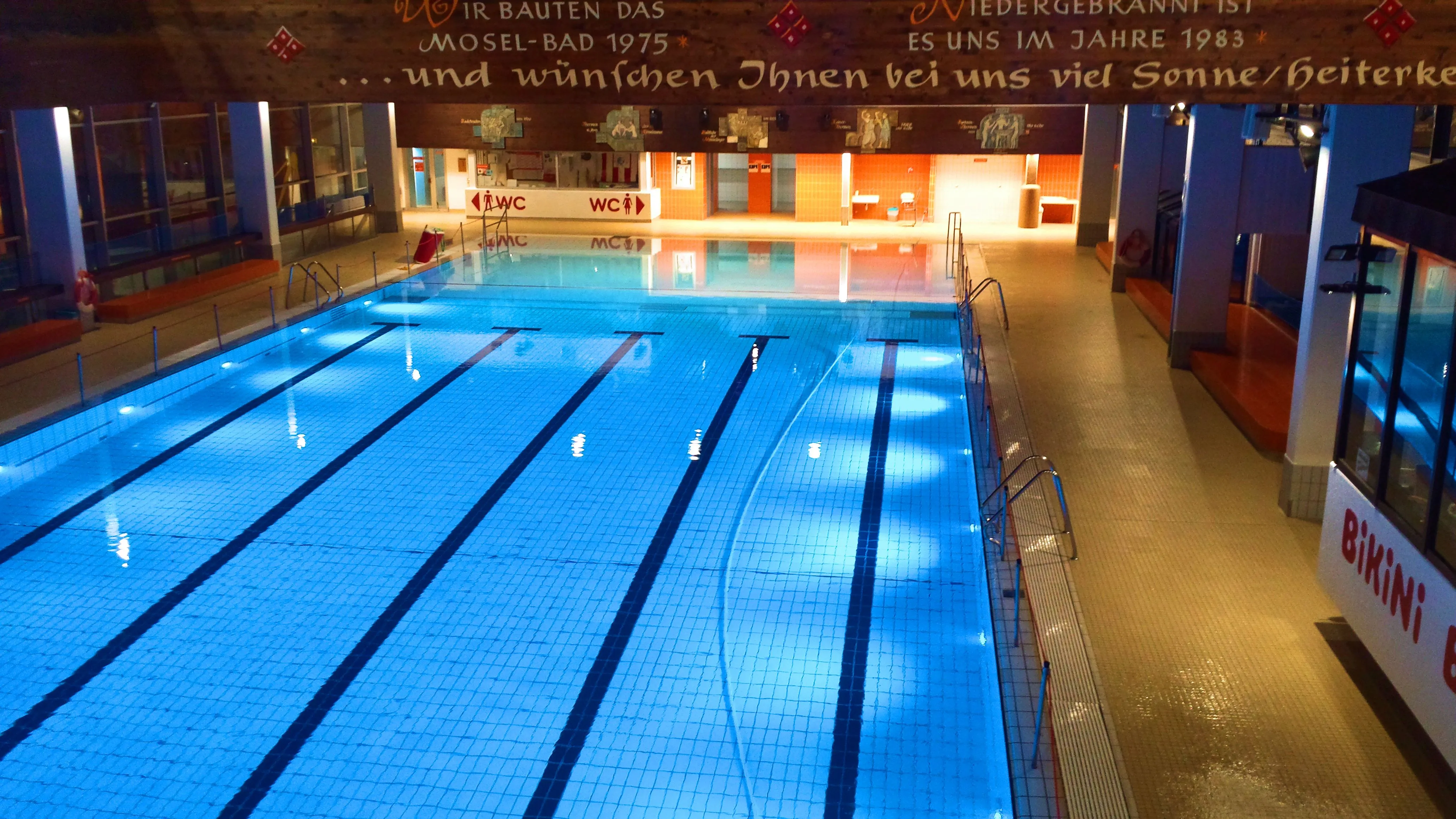 Leeres Schwimmerbecken mit blauem Wasser und markierten Bahnen in einer Schwimmhalle mit bemaltem Holzbalken
