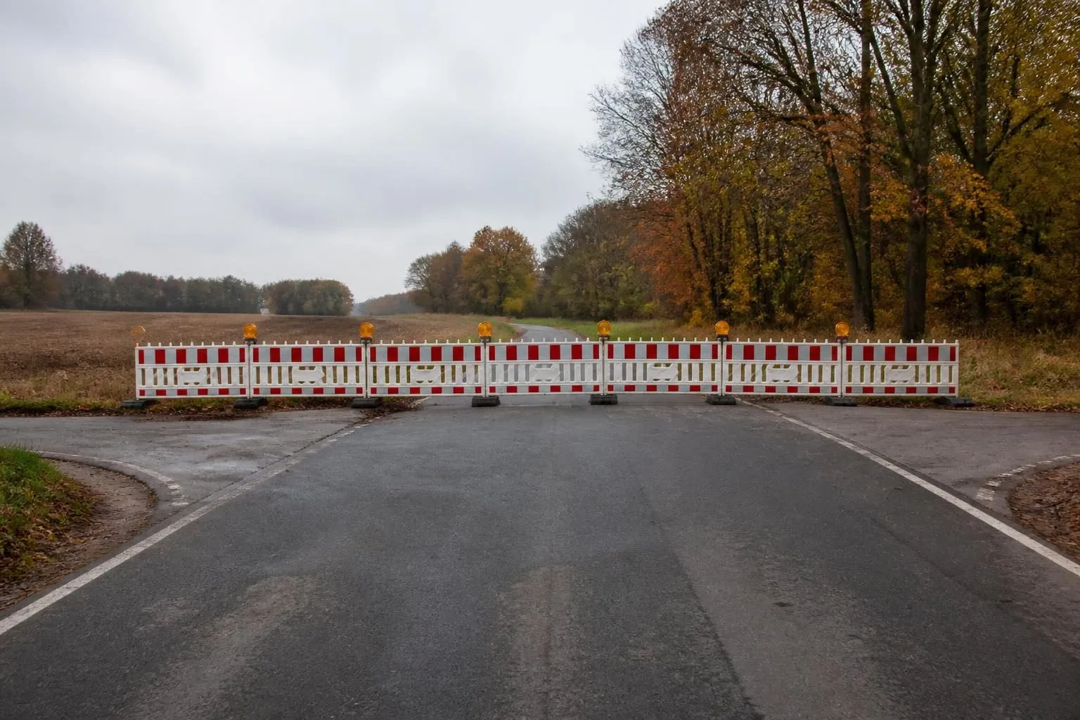Rot-weiße Absperrgitter mit gelben Warnleuchten quer über einer leeren Landstraße, Felder und Herbstlaub im Hintergrund