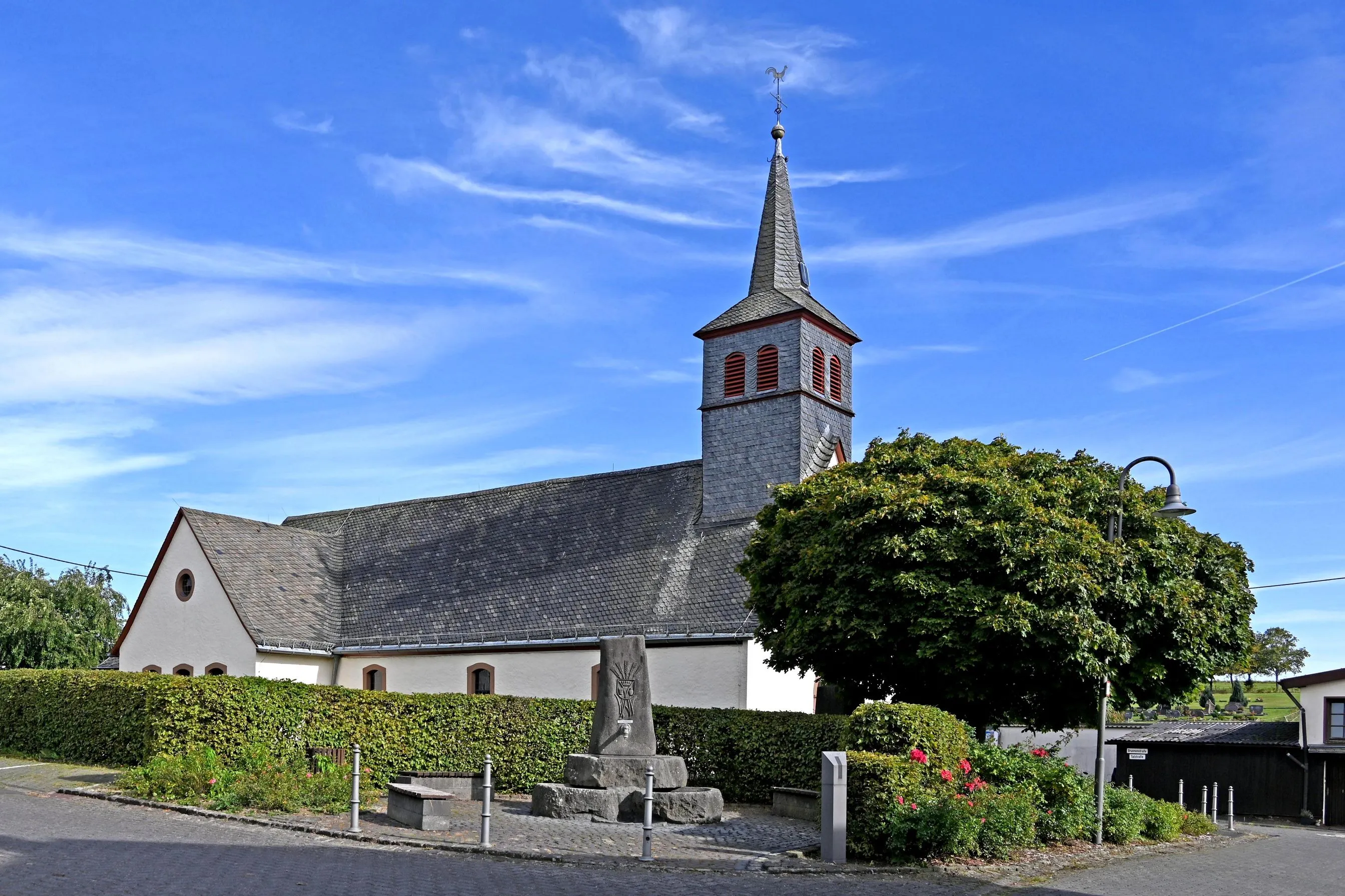 Weiß verputzte Dorfkirche mit Schieferdach hinter einer Hecke, davor ein steinerner Brunnen und eine gepflasterte Straße
