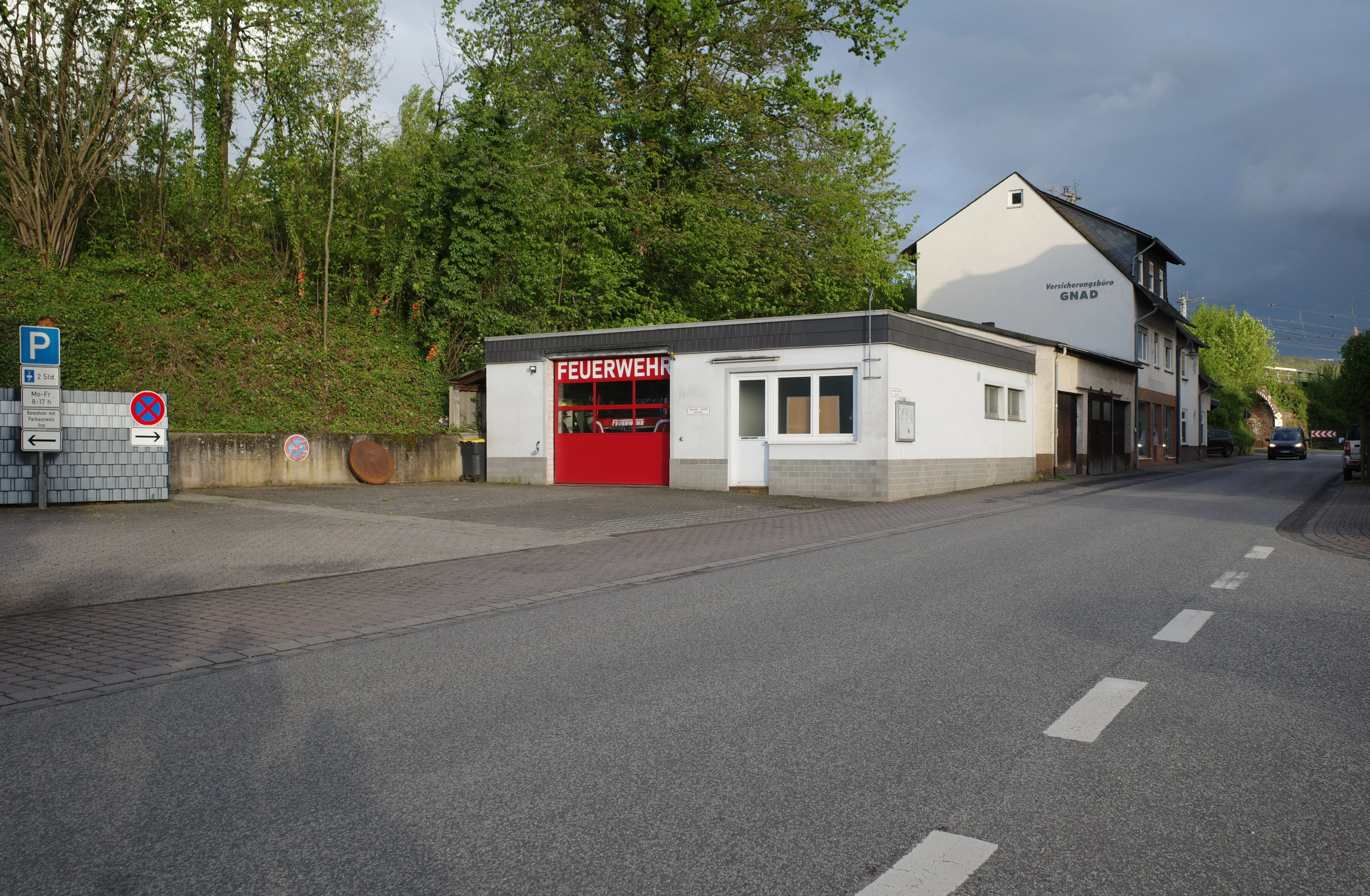 Gerätehaus der Freiwilligen Feuerwehr Bullay an der Ortsdurchfahrt, rotes Rolltor, bewachsener Hang dahinter