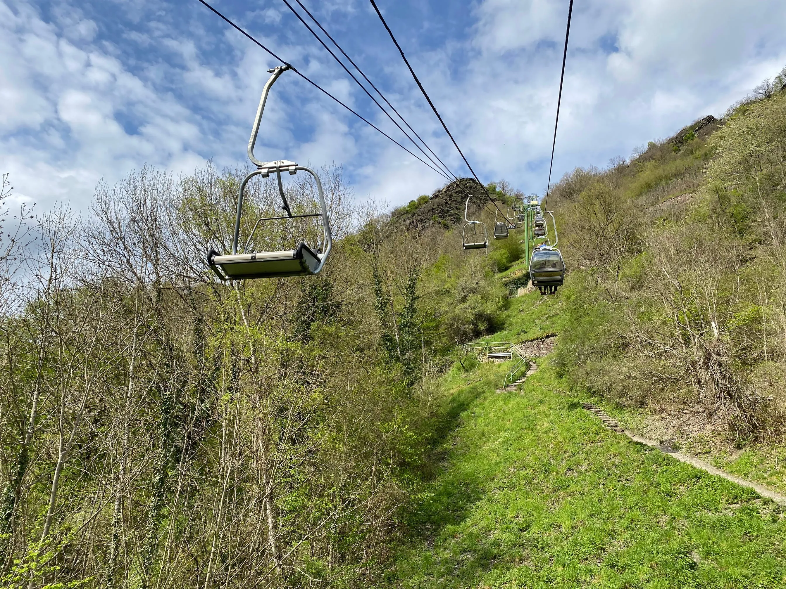 Sesselbahn über einem grünen Hang, daneben ein steiler Treppenweg bergauf, im Hintergrund bewaldete Höhen