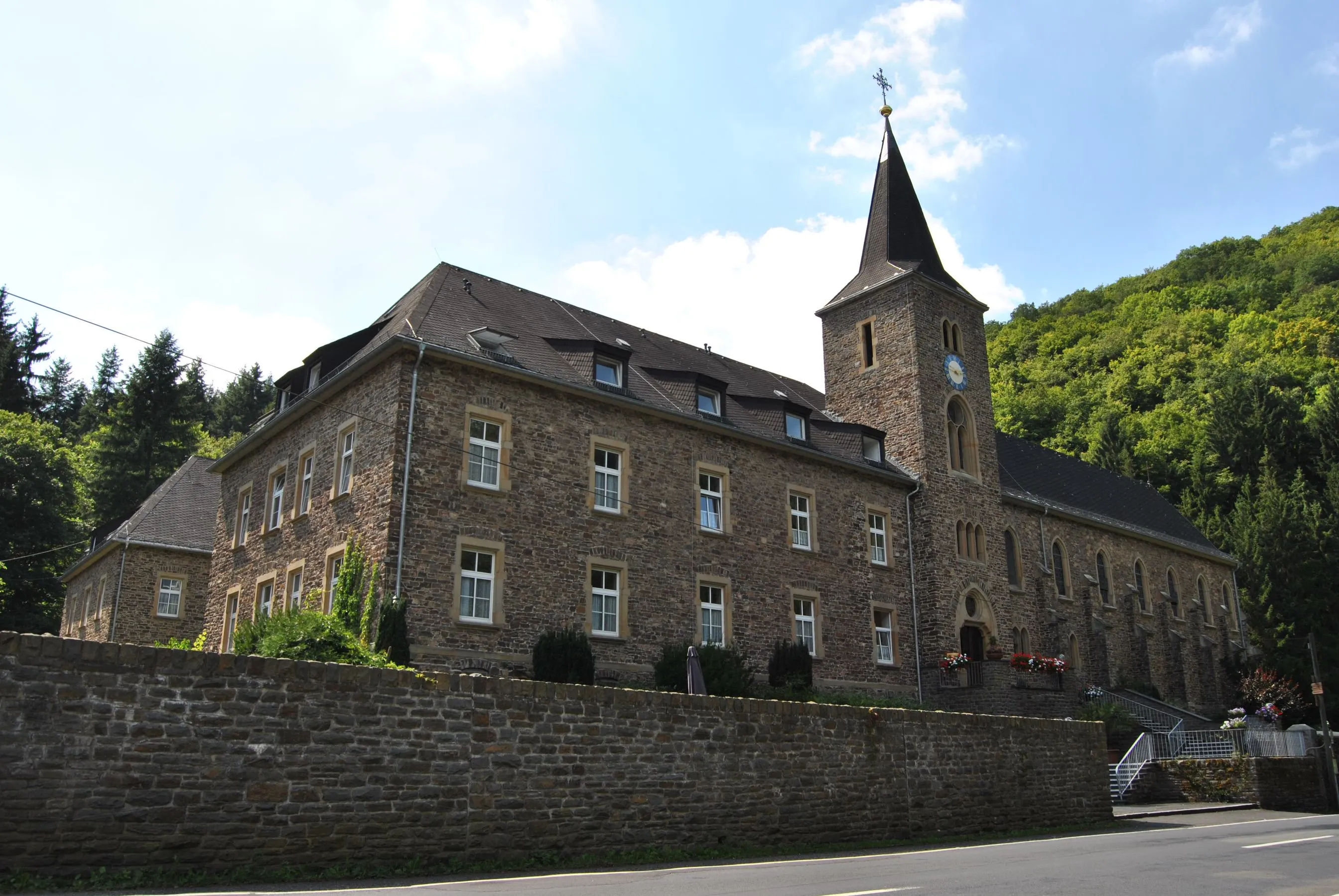 Kloster Maria Engelport im Flaumbachtal: Klostergebäude aus Bruchstein mit Kirchturm vor einem bewaldeten Hang
