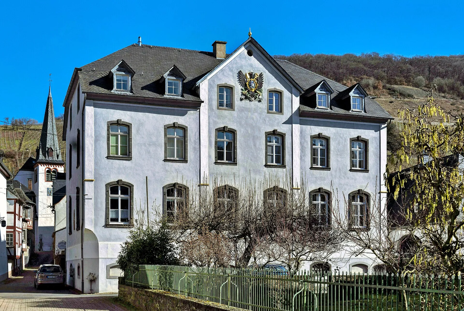 Der spätbarocke Brauweiler Hof in Mesenich mit dem Wappenadler im Giebel, links der Kirchturm, dahinter der Weinberg