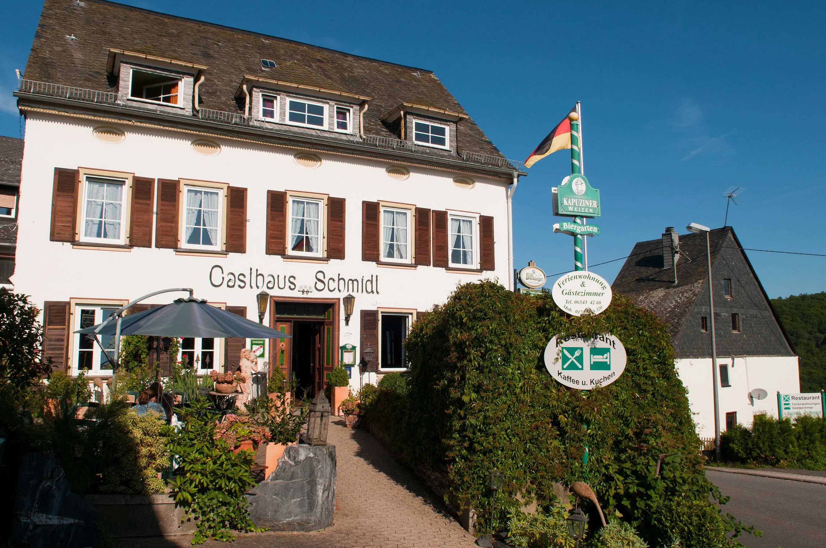 Weiß verputztes Gasthaus mit Schieferdach, Schriftzug über der Tür, Biergartenschildern am Mast und bewachsener Hecke davor