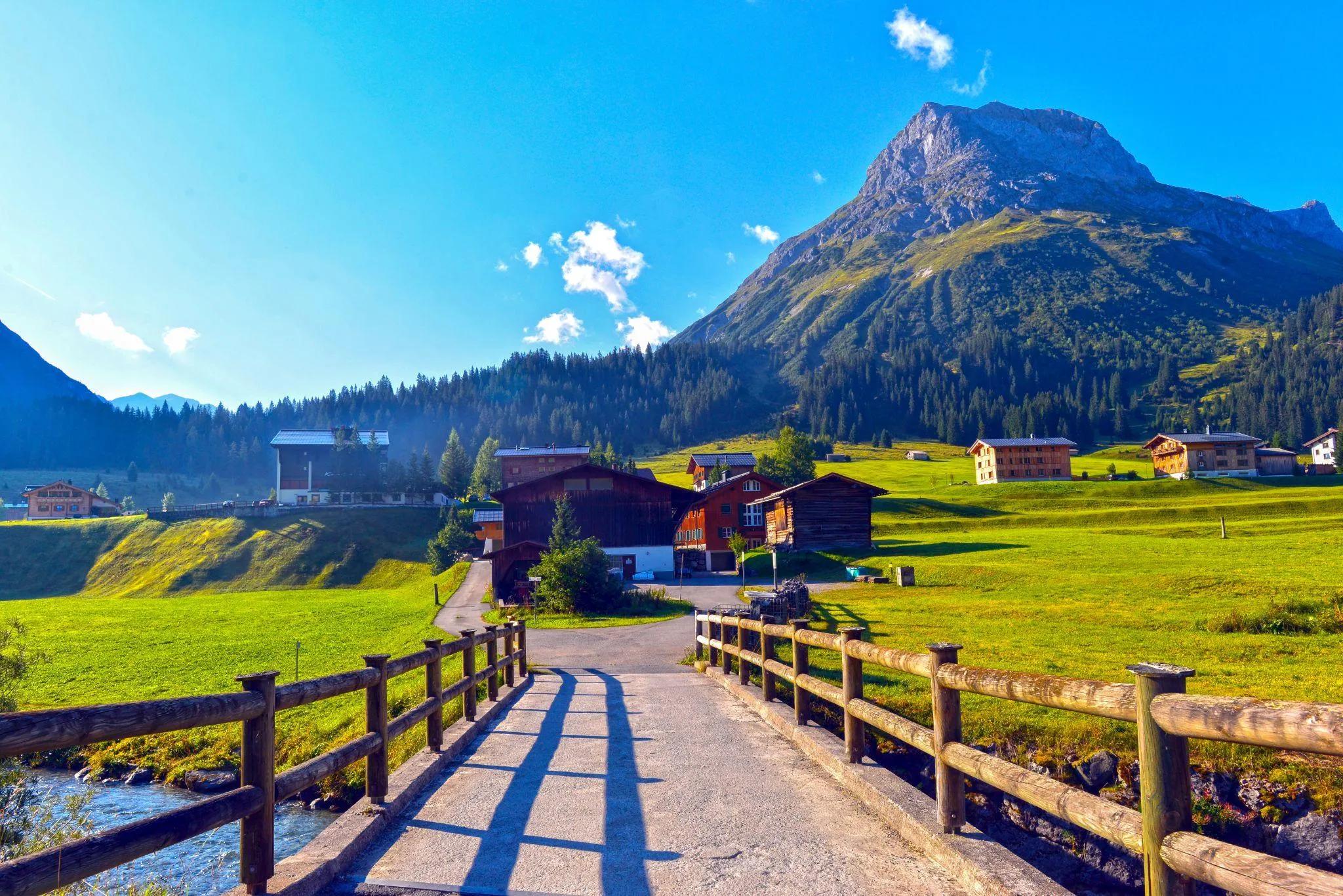 Holzbrücke über einen Bergbach, dahinter Holzhäuser auf grünen Wiesen und ein felsiger Gipfel unter blauem Himmel