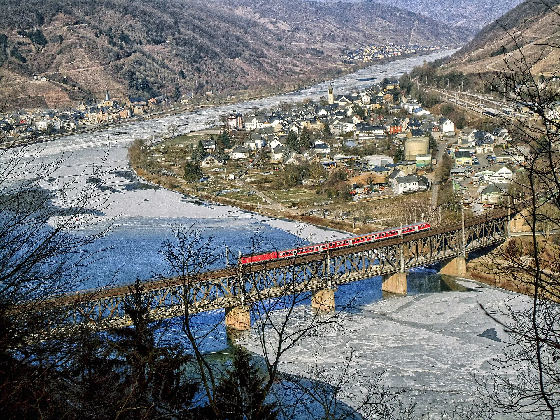 Stahlfachwerkbrücke über die winterliche Mosel mit einem roten Regionalzug, dahinter der Ort Bullay im Tal