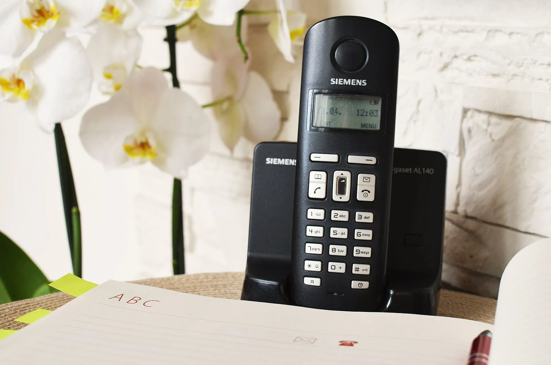 Schnurloses Telefon in seiner Ladestation auf einem Tisch, daneben ein aufgeschlagenes Adressbuch und eine weiße Orchidee