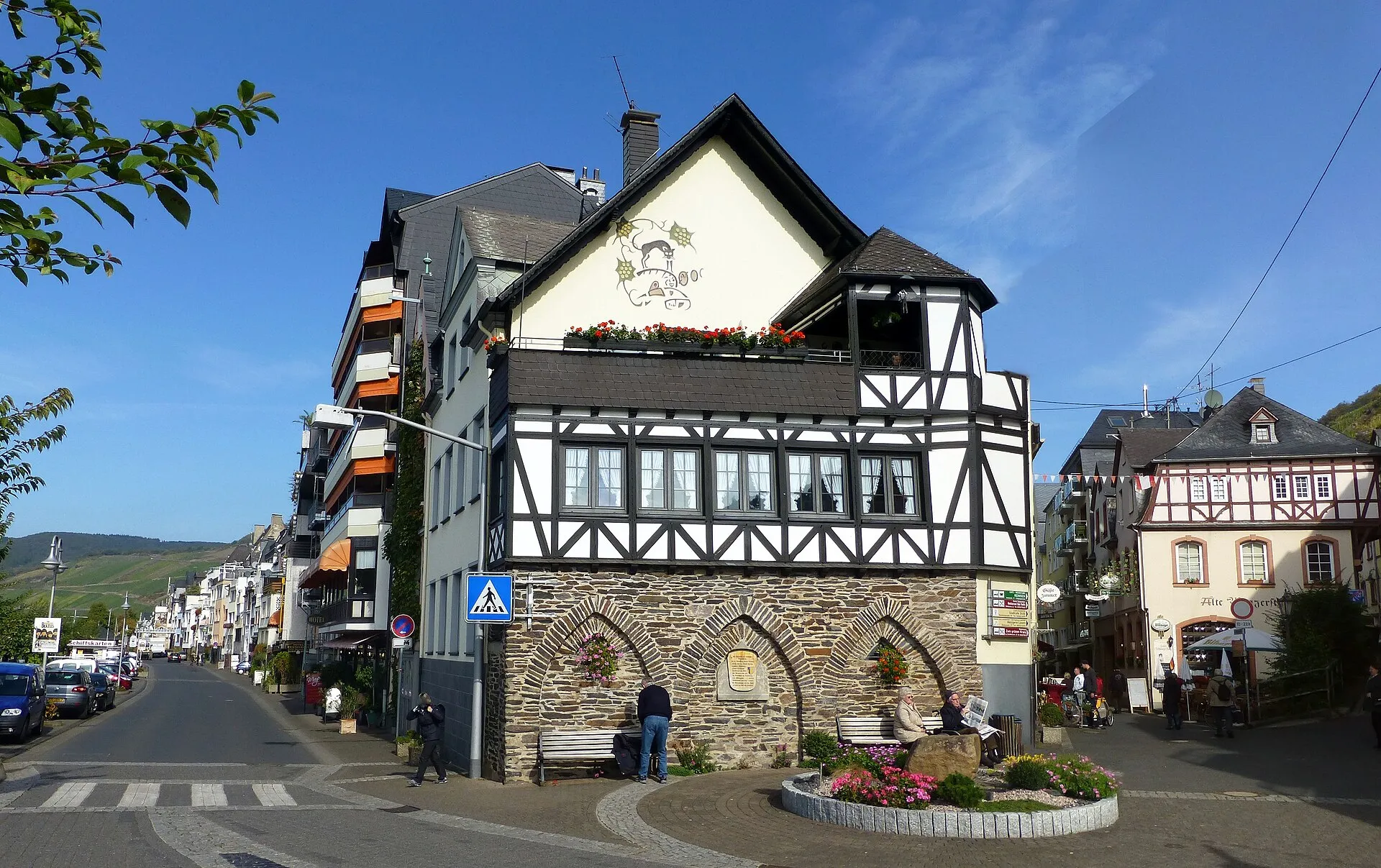 Fachwerkhaus mit Schiefersockel und Rundbögen an einer Straßenecke, daneben eine Geschäftszeile und ein Wimpelband