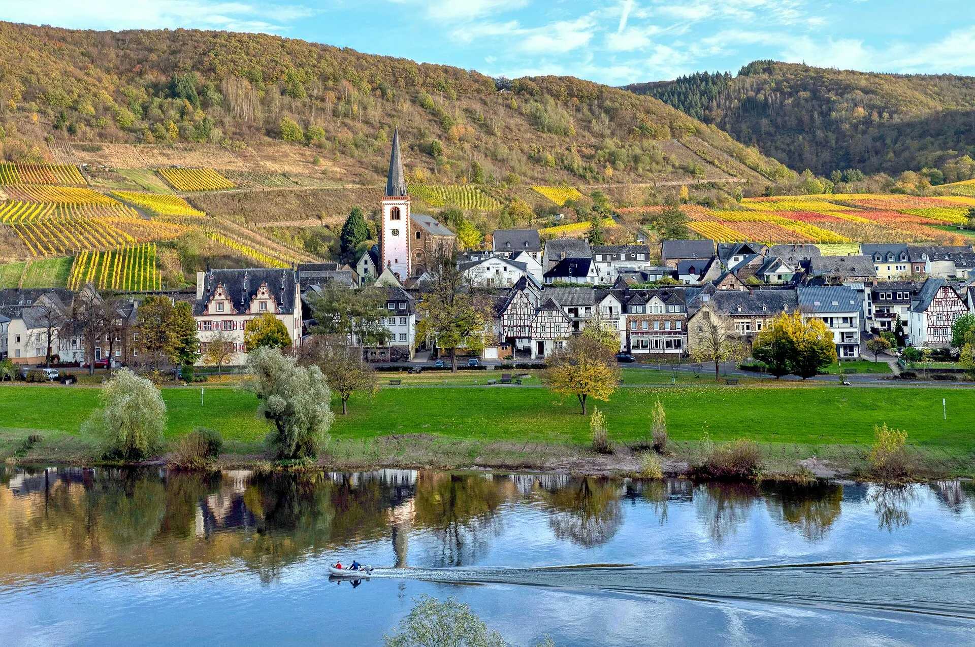 Blick über die Mosel auf Bruttig: Kirchturm, Häuserzeile am Ufer, dahinter herbstlich gefärbte Weinberge