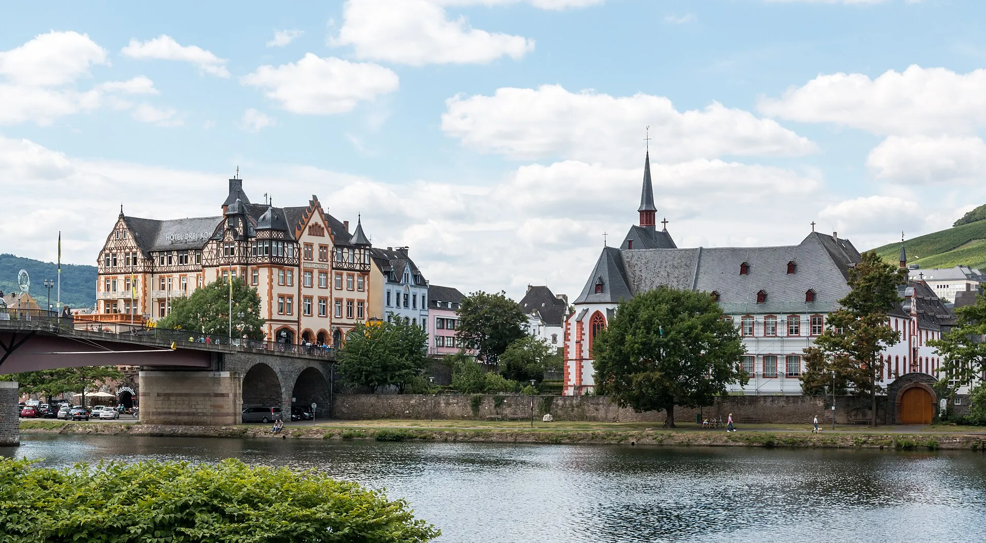 Die Uferzeile von Kues an der Mosel mit dem Sankt-Nikolaus-Hospital, der Moselbrücke und den Weinbergen dahinter