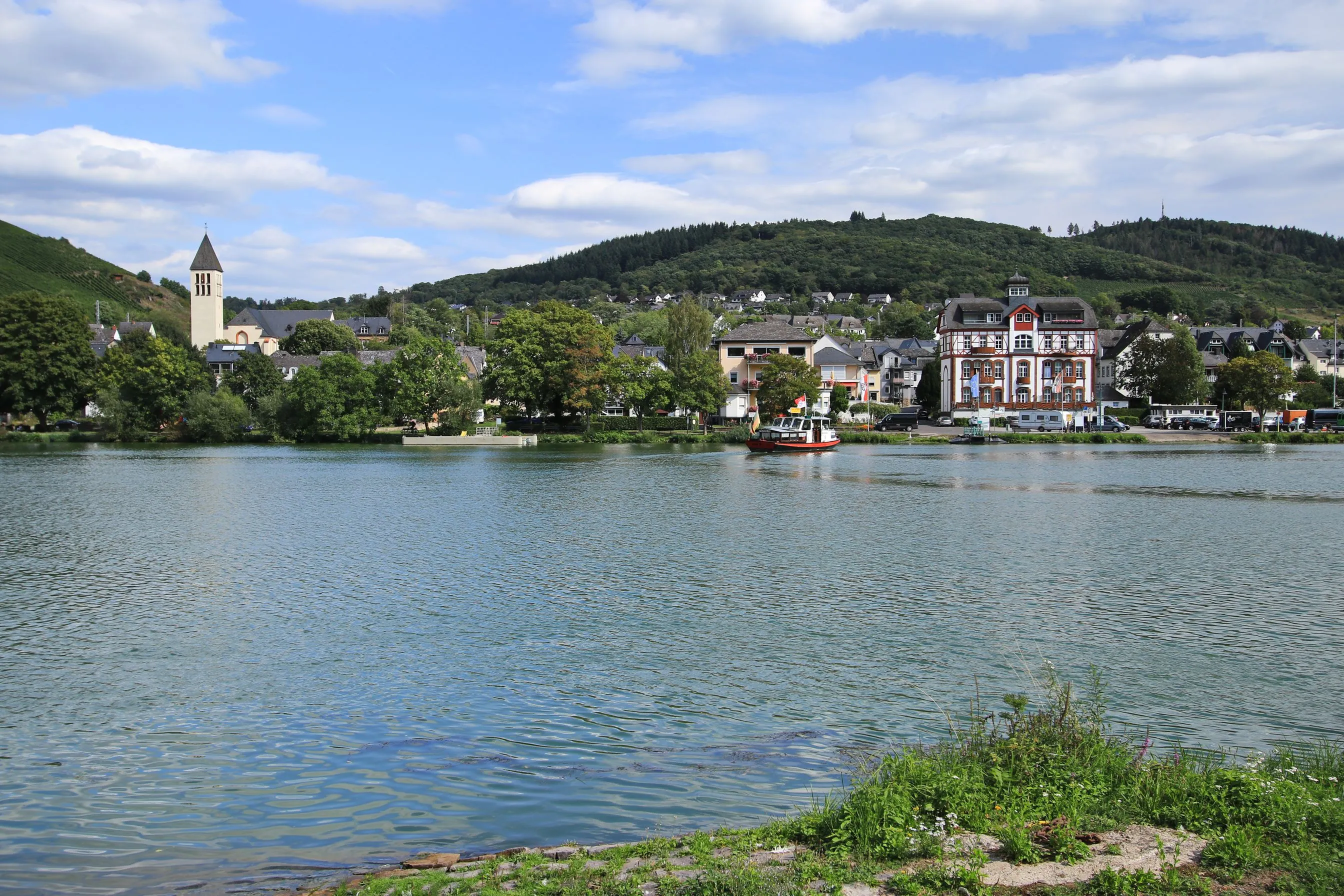 Blick über die Mosel auf Bullay mit Kirchturm, einem großen Fachwerkhaus am Ufer und einer kleinen Fähre auf dem Wasser