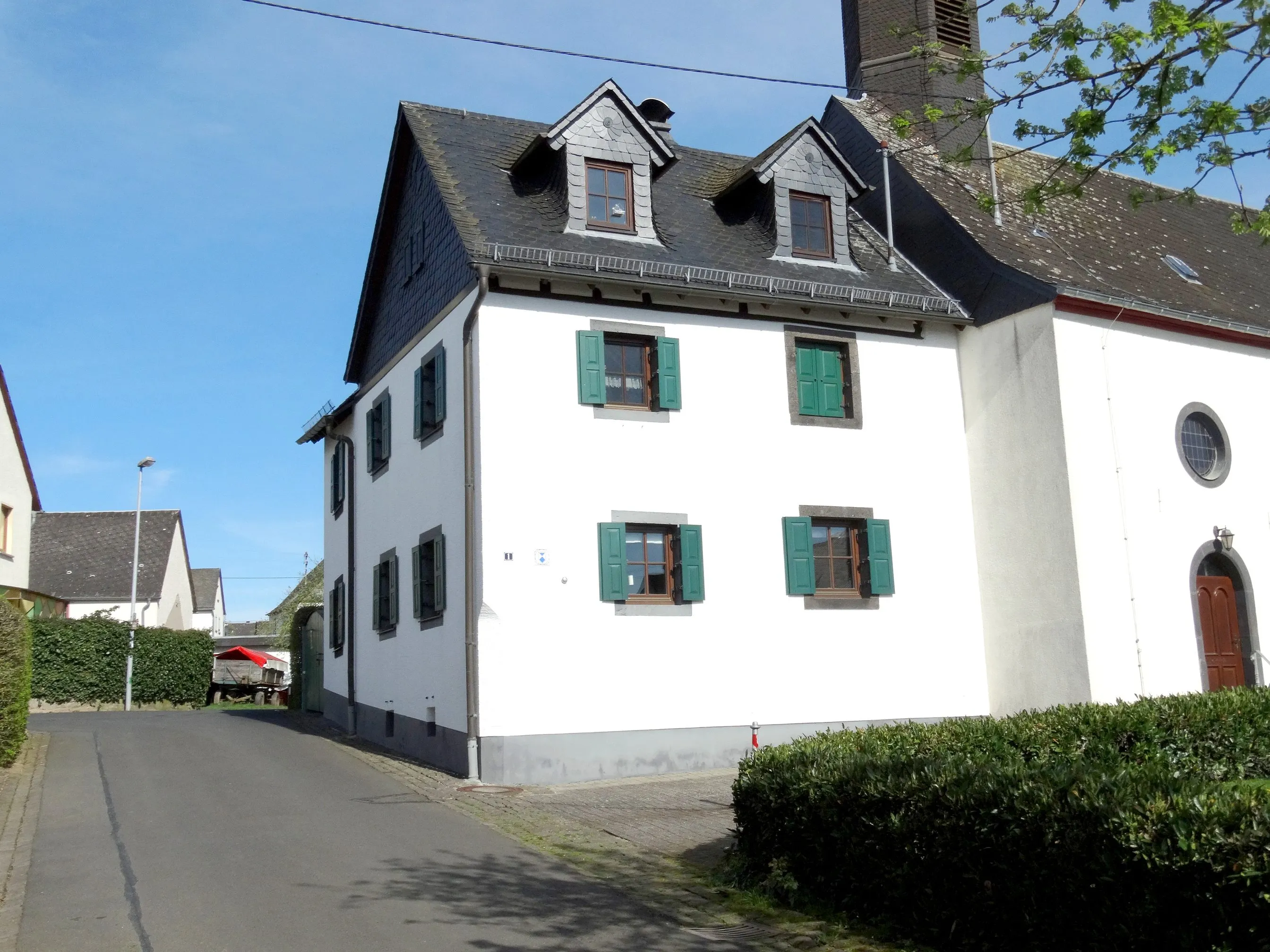 Weiß verputztes Haus mit grünen Fensterläden und Schieferdach in Greimersburg, an die Kapelle gebaut, davor die Dorfstraße