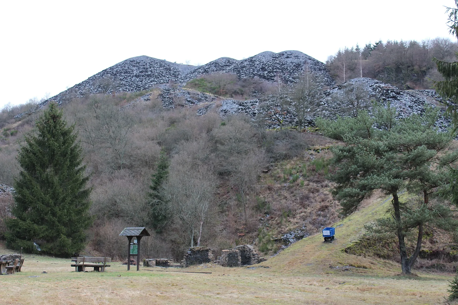 Graue Schieferhalden über einem Wiesengrund, davor Infotafel, Bänke, Reste einer Schiefermauer und ein blauer Förderwagen