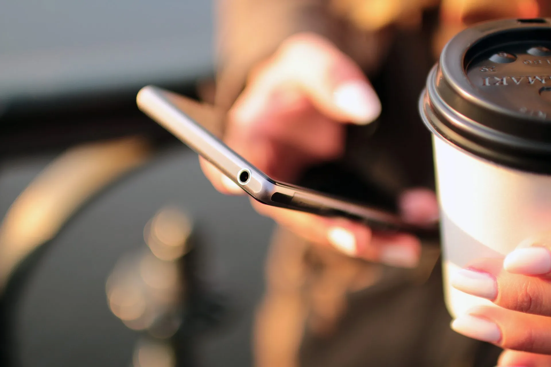 Hand hält ein Smartphone, in der anderen Hand ein Kaffeebecher, der Hintergrund ist unscharf