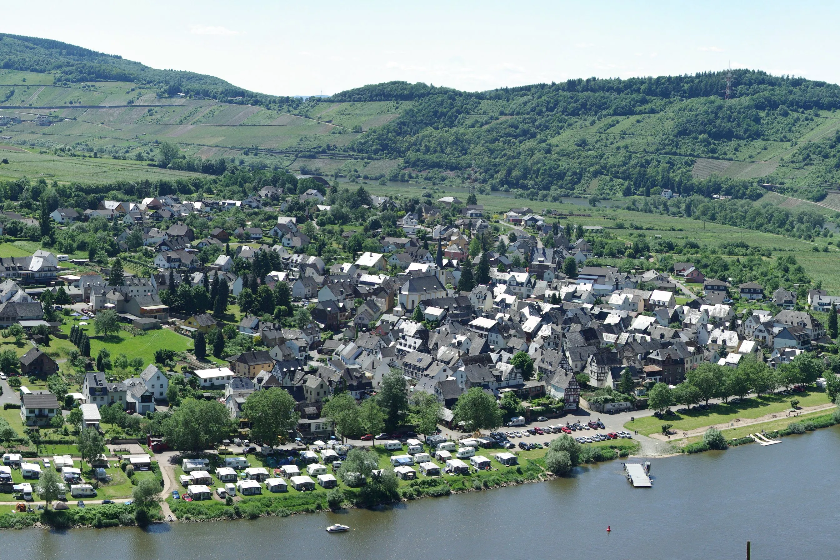 Blick von oben auf Pünderich an der Mosel mit Ortskern, Campingplatz am Ufer und Weinbergen an den Hängen