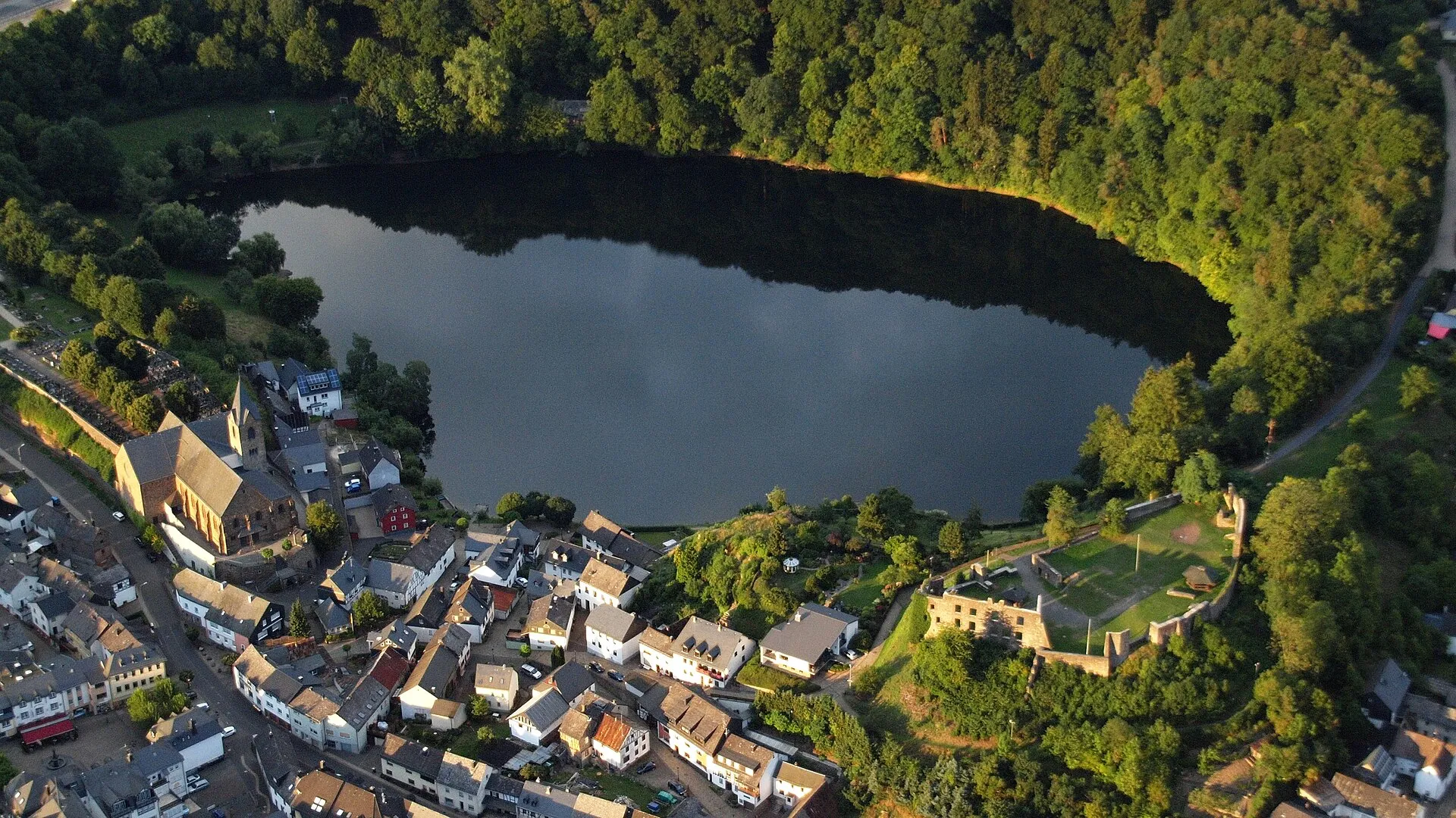 Luftbild eines dunklen runden Kratersees, links am Ufer Häuser und eine Kirche, rechts eine Burgruine auf dem Wall