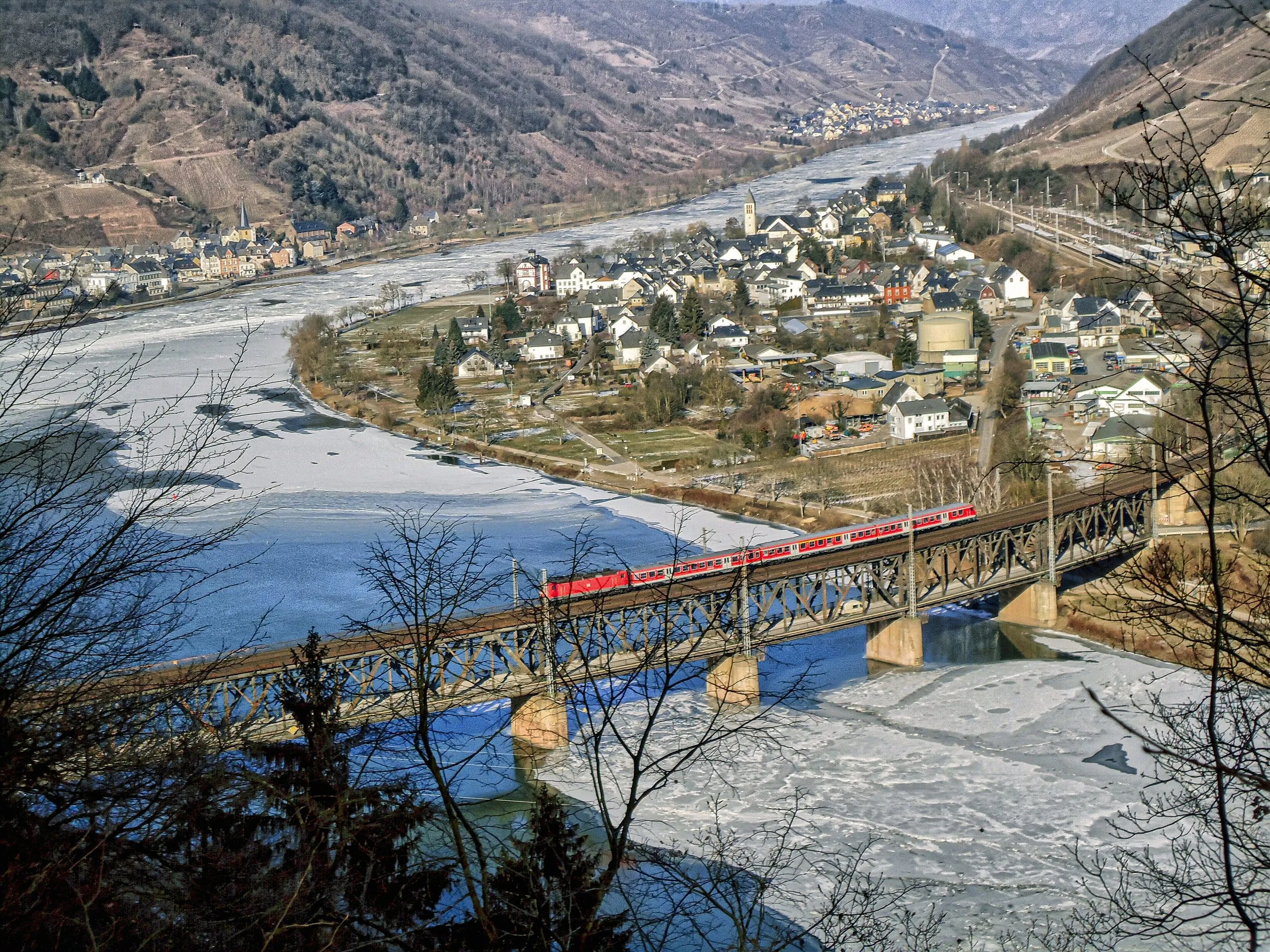 Bullay an der winterlichen Mosel von oben, Eisschollen auf dem Fluss, ein roter Zug auf der Doppelstockbrücke