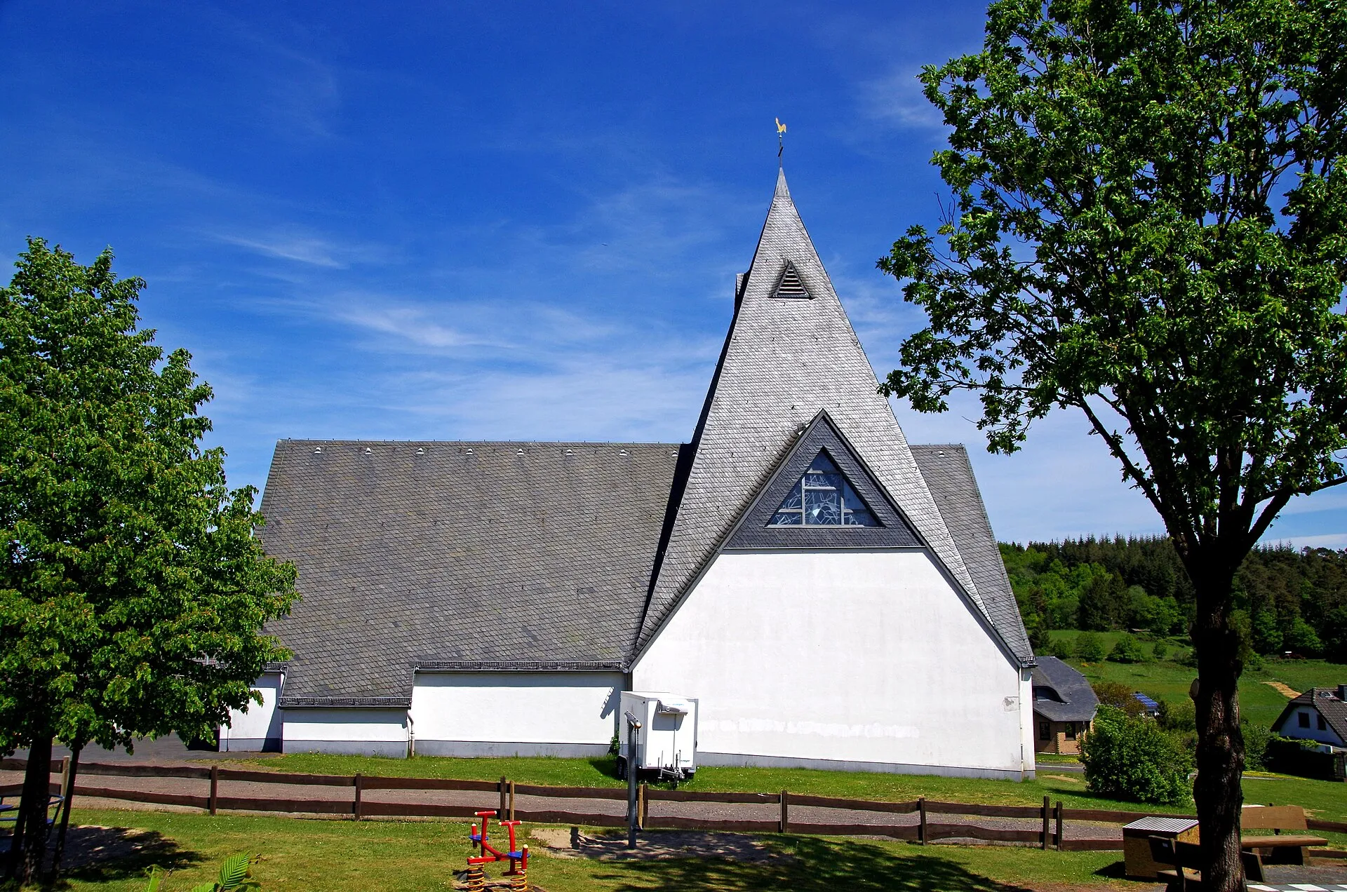 Kirche St. Josef in Laubach von Westen: weißer Giebel unter einem steilen Schieferdach, davor Wiese und Spielgeräte