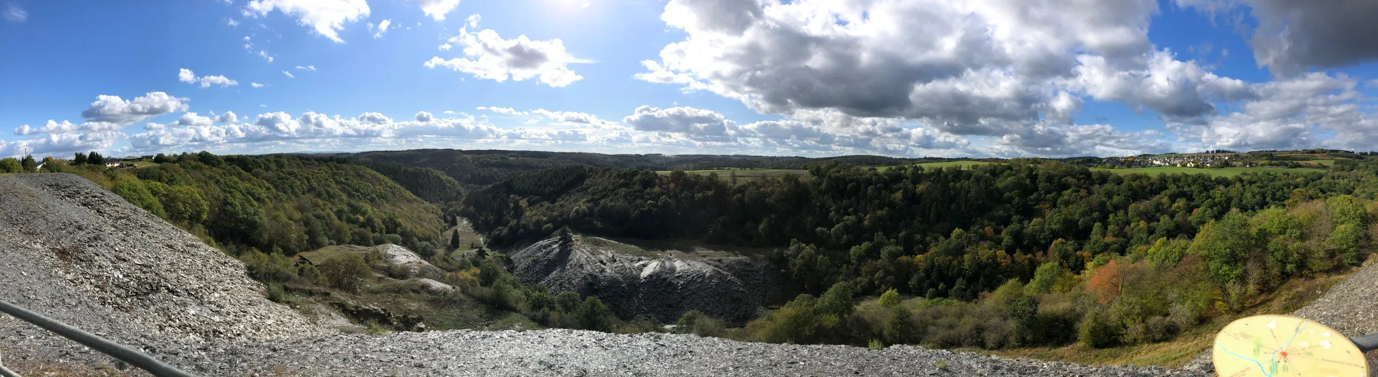 Weiter Blick über ein bewaldetes Tal bei Leienkaul, am Hang eine helle Halde aus Schiefergestein, darüber Wolkenhimmel