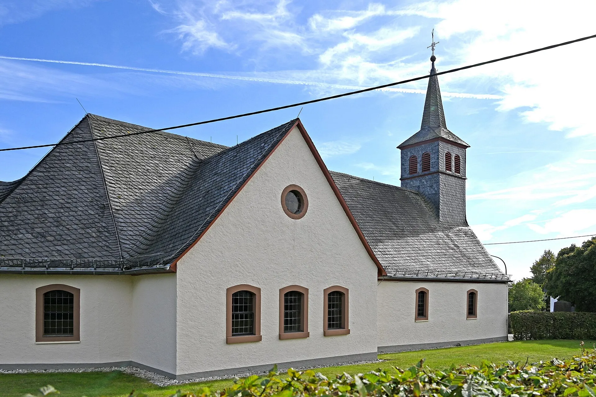 Weiß verputzte Dorfkirche mit Schieferdach und schlankem Turm, davor Rasen und geschnittene Hecke unter blauem Himmel