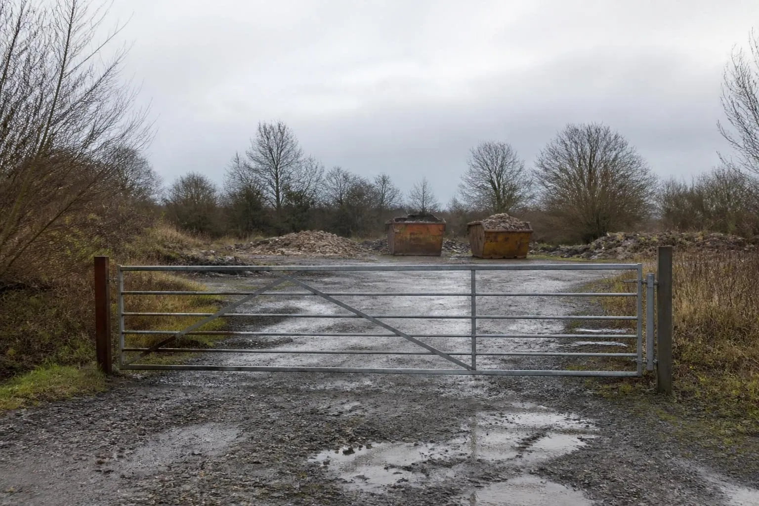 Geschlossenes Metalltor vor einem nassen Schotterplatz, dahinter zwei Absetzcontainer mit Bauschutt und kahle Bäume