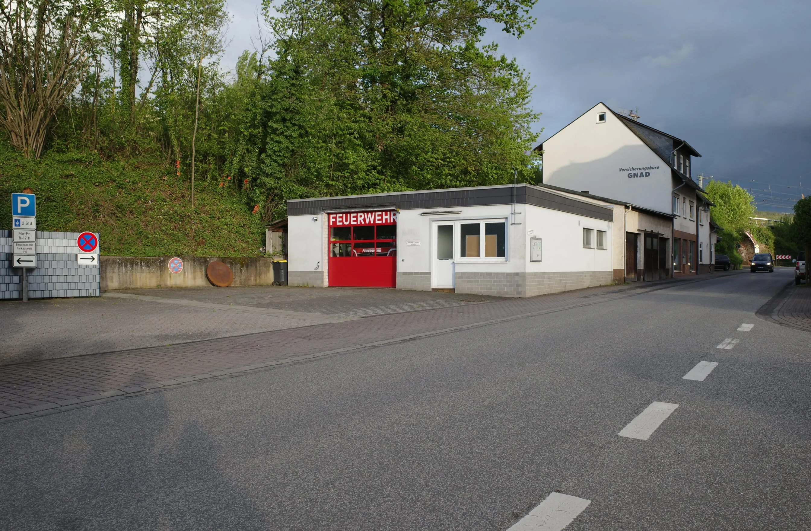 Gerätehaus der Freiwilligen Feuerwehr Bullay an der Ortsdurchfahrt, rotes Rolltor, bewachsener Hang dahinter