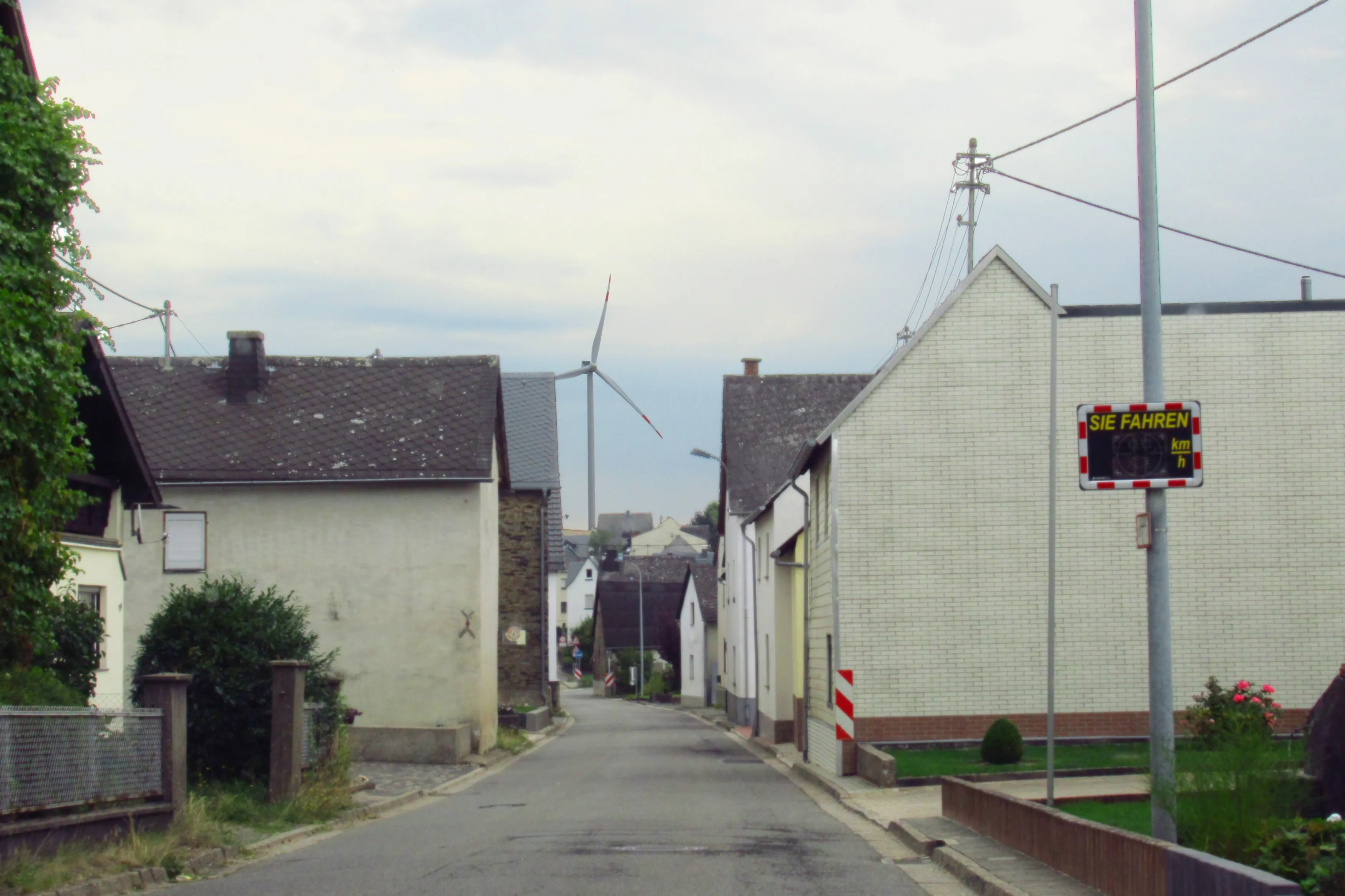 Dorfstraße mit verputzten Häusern und Schieferdächern, dahinter ein Windrad über den Feldern der Vordereifel