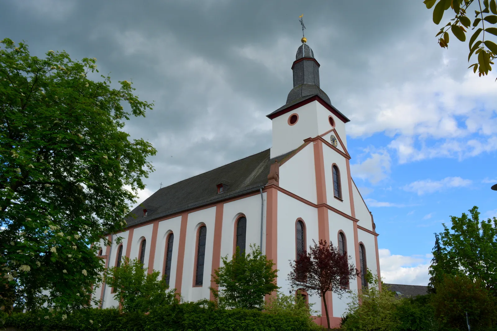 Weiß-rot gefasste Hallenkirche mit schiefergedecktem Turmhelm, davor Bäume und Hecken unter wolkigem Himmel