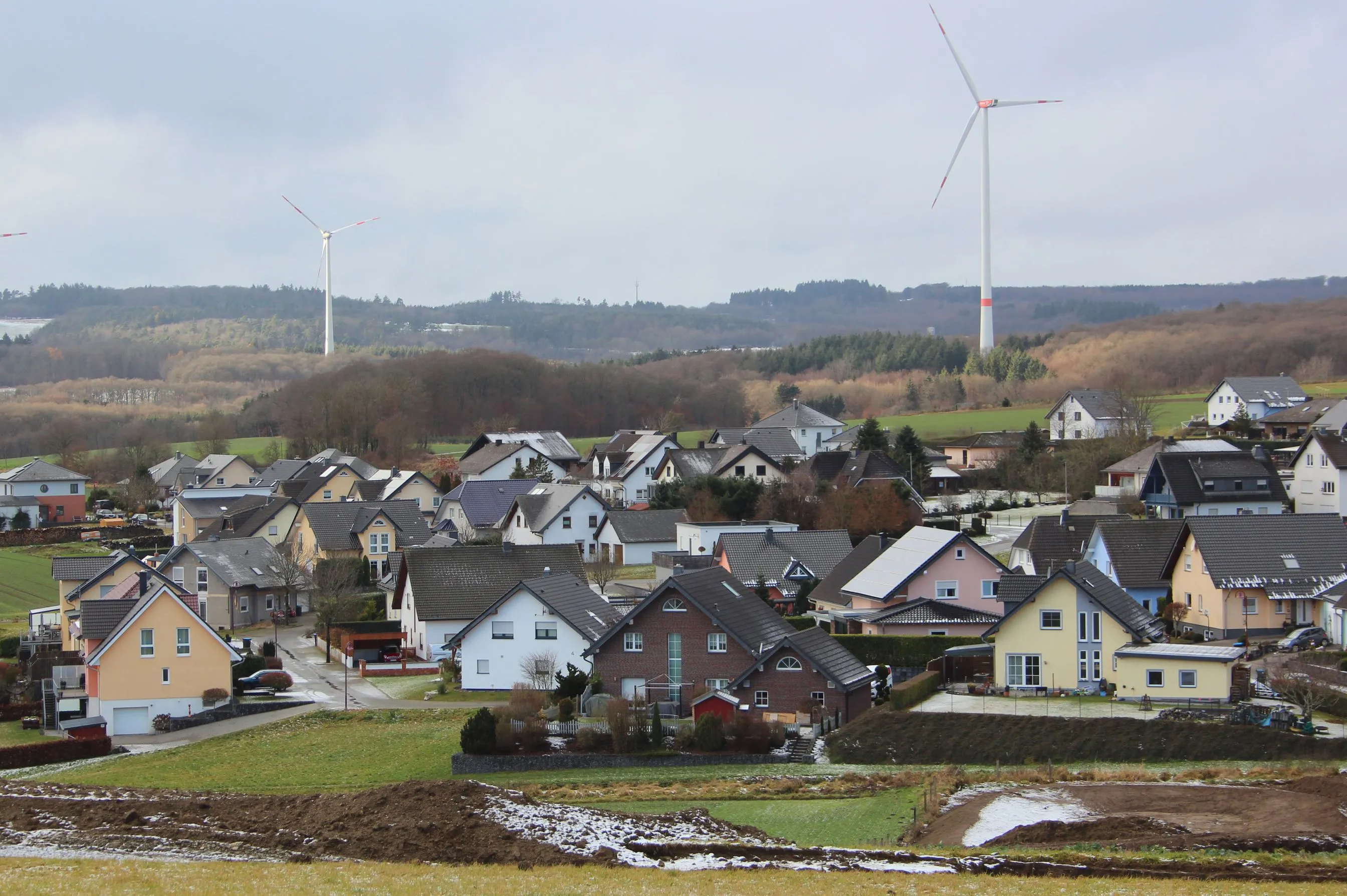 Blick über das Eifeldorf Hambuch mit Wohnhäusern, dahinter bewaldete Höhenrücken und zwei Windräder bei Winterwetter