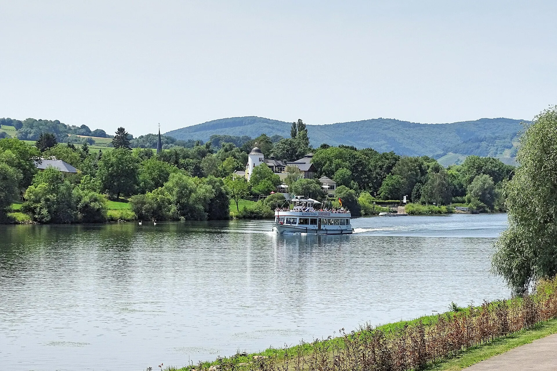 Ein Ausflugsschiff fährt auf der Mosel an einem Uferort vorbei, dahinter bewaldete Hänge und ein Kirchturm