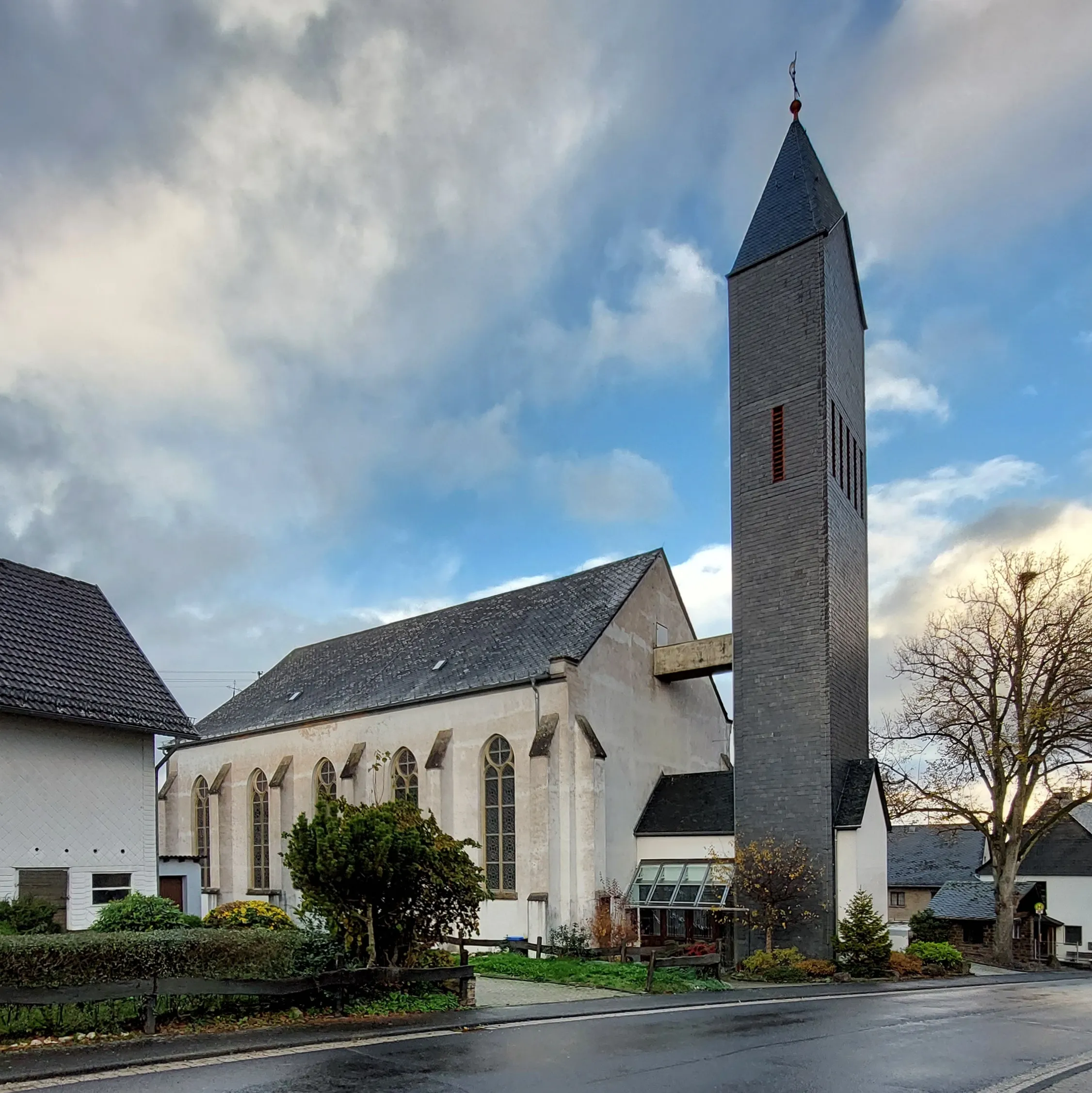 Katholische Dorfkirche mit hohem schiefergedecktem Glockenturm an einer Straße, daneben Wohnhäuser und ein kahler Baum