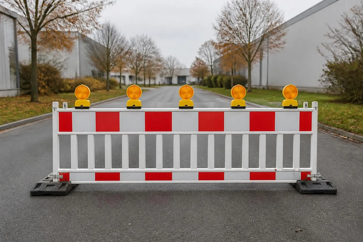 Rot-weiße Absperrschranke mit gelben Warnleuchten quer über einer leeren Asphaltstraße zwischen Hallen, kahle Bäume dahinter