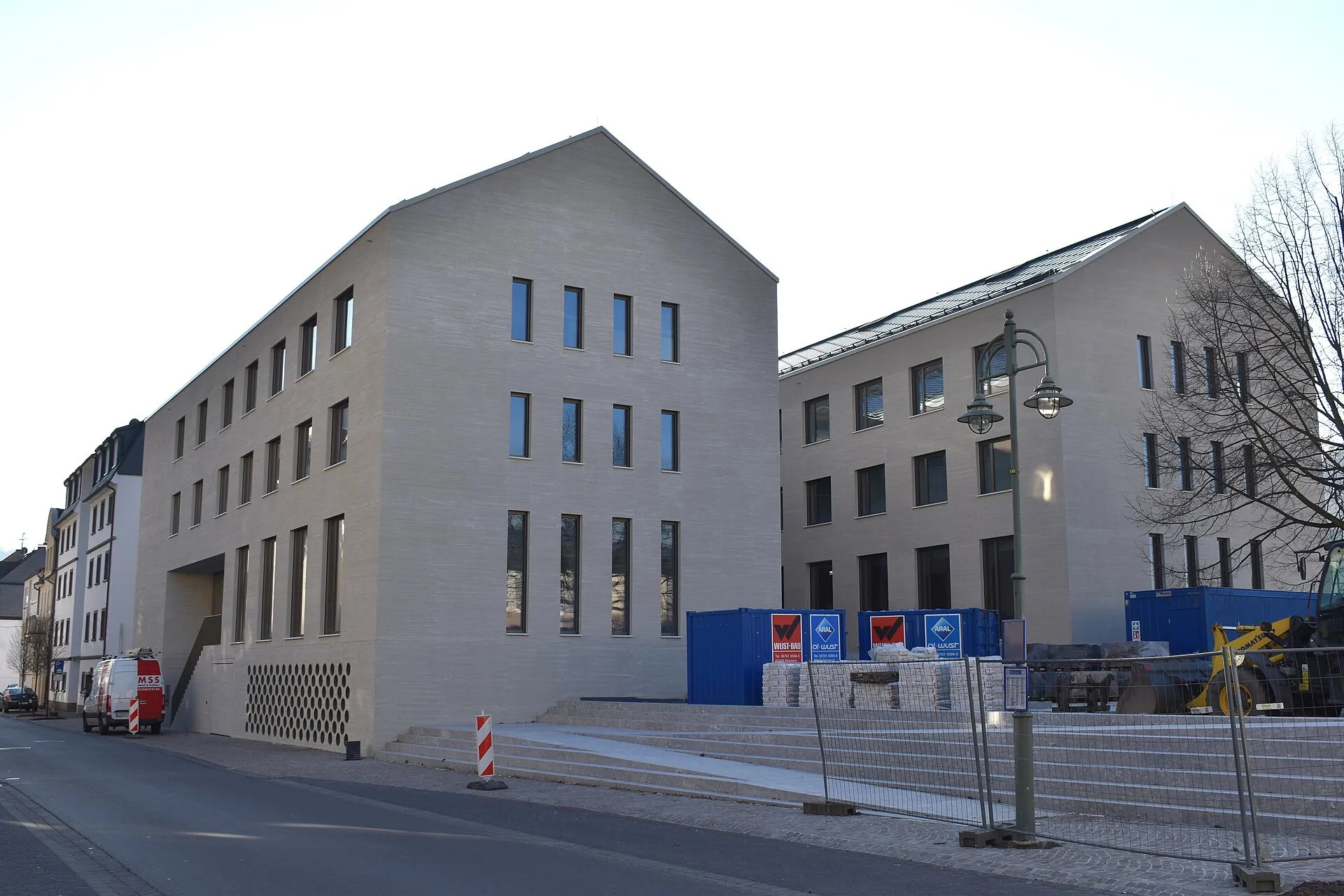 Heller dreigeschossiger Verwaltungsneubau mit Satteldach an einer Straße, davor Bauzaun, Baucontainer und neue Treppenstufen
