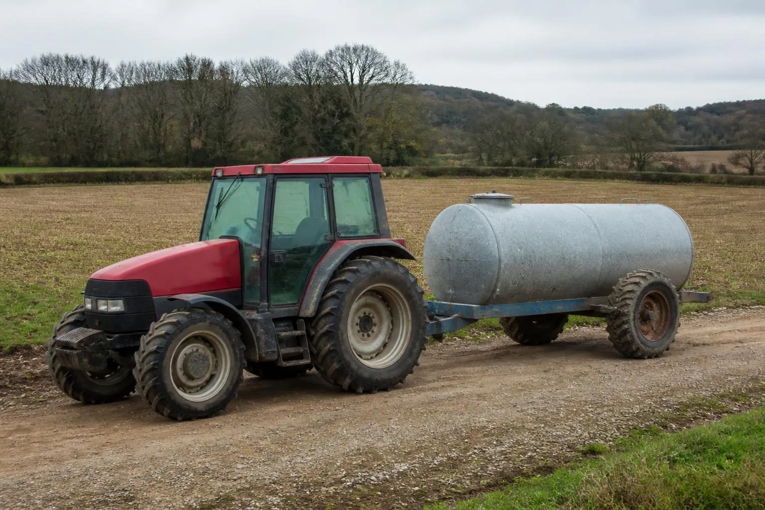 Traktor mit angehängtem verzinktem Wassertank auf einem Schotterweg am Rand eines abgeernteten Feldes