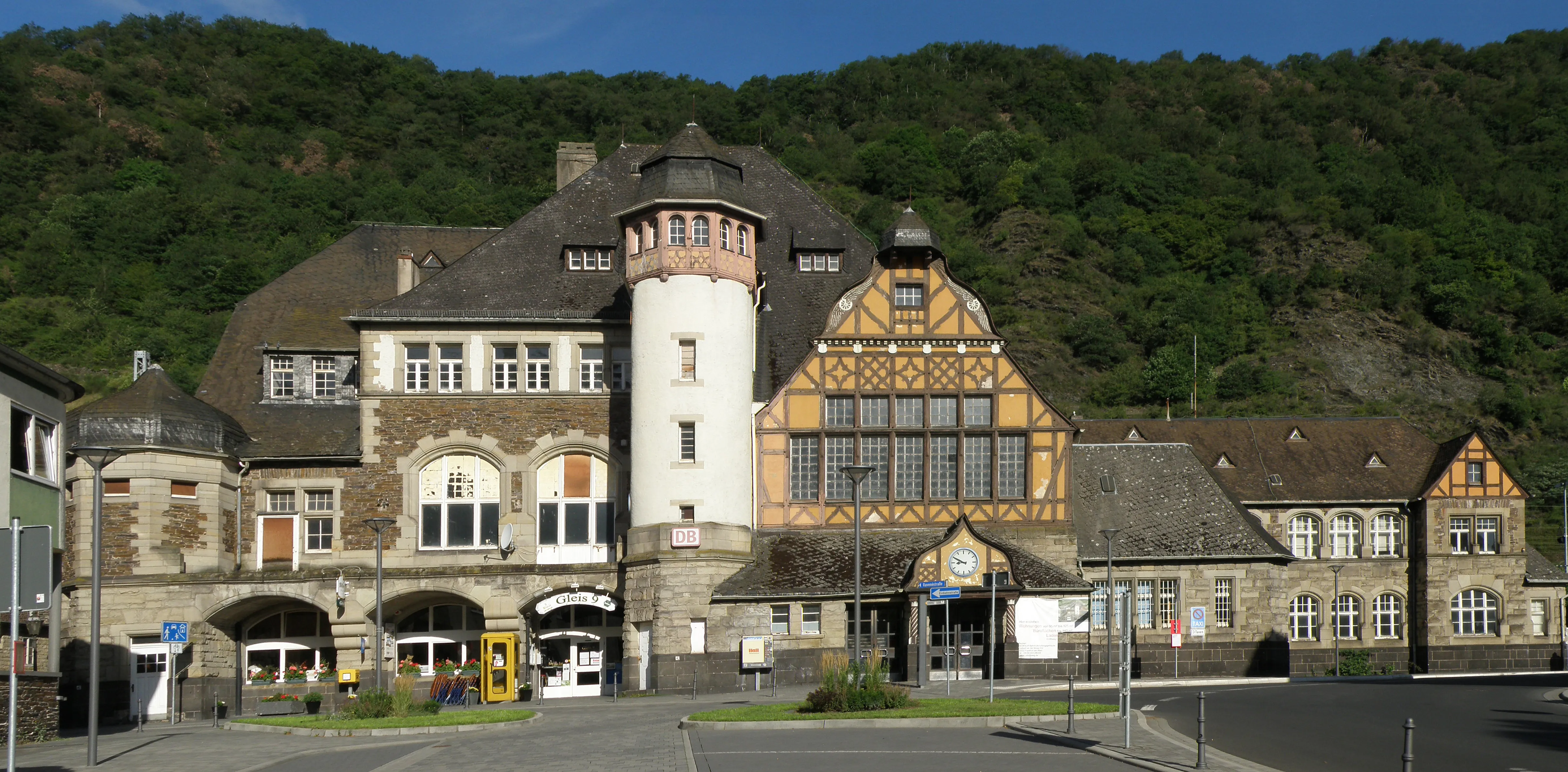 Bahnhofsgebäude von Cochem mit Fachwerkgiebel, Uhrenturm und Schieferdach, davor der leere gepflasterte Vorplatz