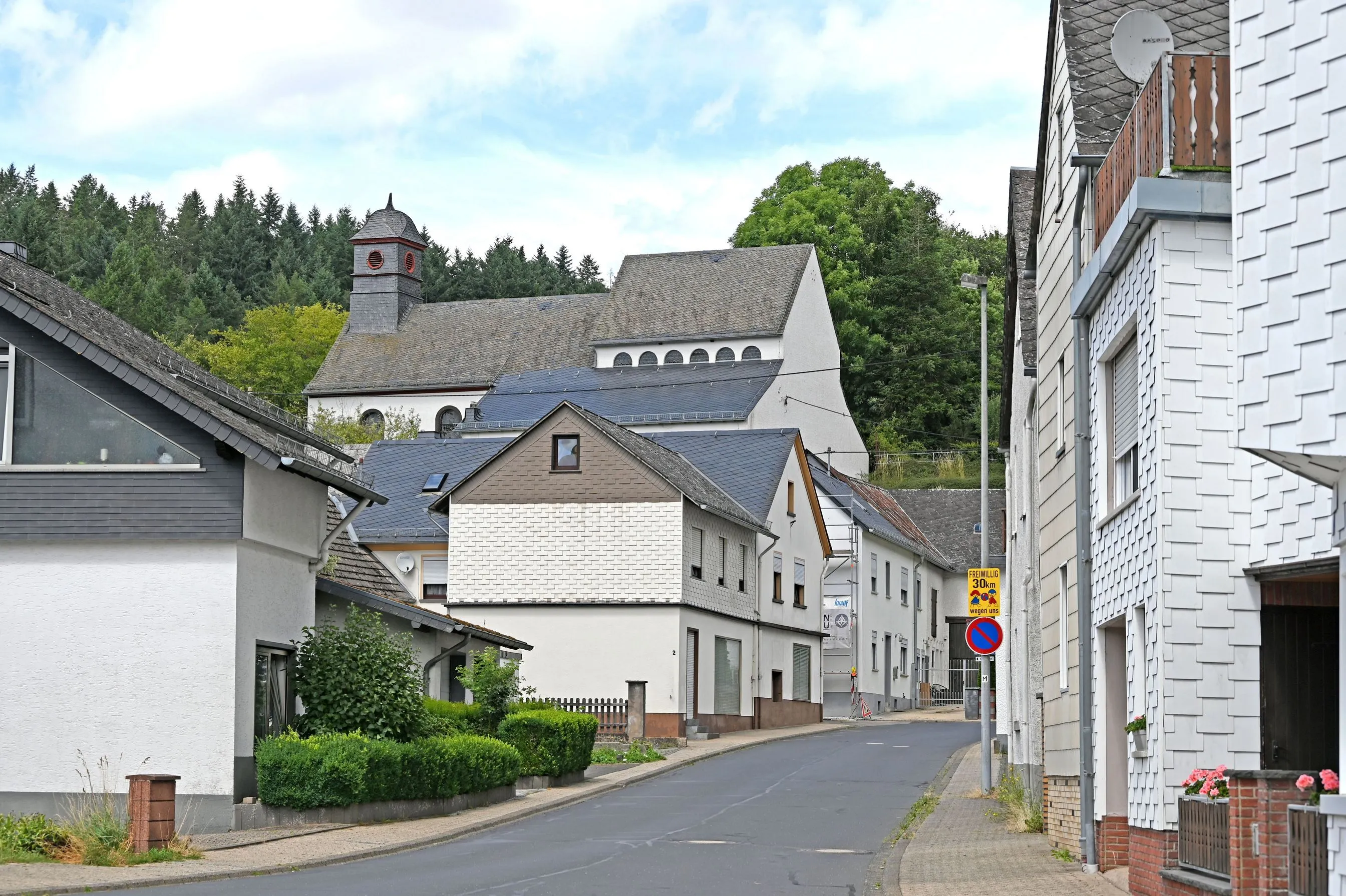 Ortsstraße in Urmersbach mit verschieferten und verputzten Häusern, darüber am Hang eine Kirche und Nadelwald