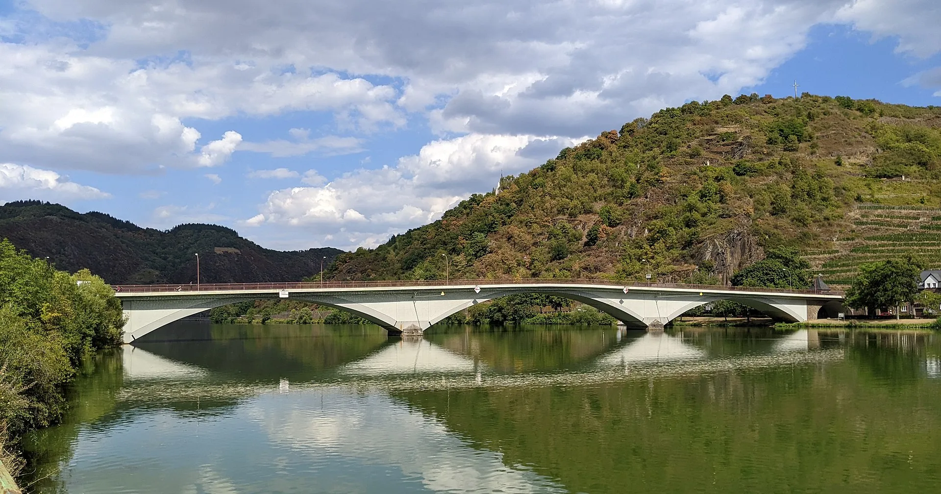 Weiße Bogenbrücke über die Mosel bei Treis-Karden, die Bögen spiegeln sich im ruhigen Wasser, dahinter ein bewaldeter Hang